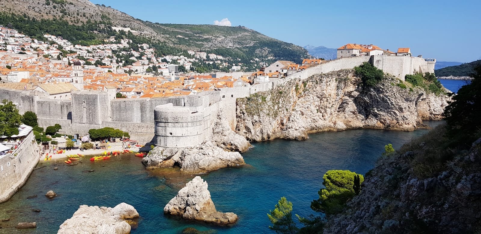 Vista panorámica de las murallas de Dubrovnik, tejados naranjas y acantilados sobre el azul del Mar Adriático en un día soleado y despejado.