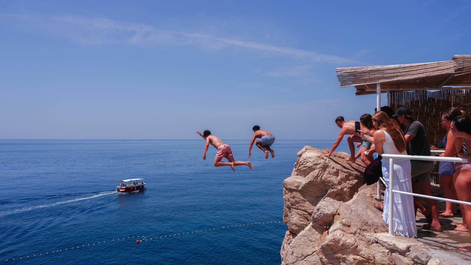 Des personnes sautent d'une terrasse de bar dans les eaux bleues et cristallines de la mer Adriatique, avec d'autres spectateurs et un bateau en arrière-plan.