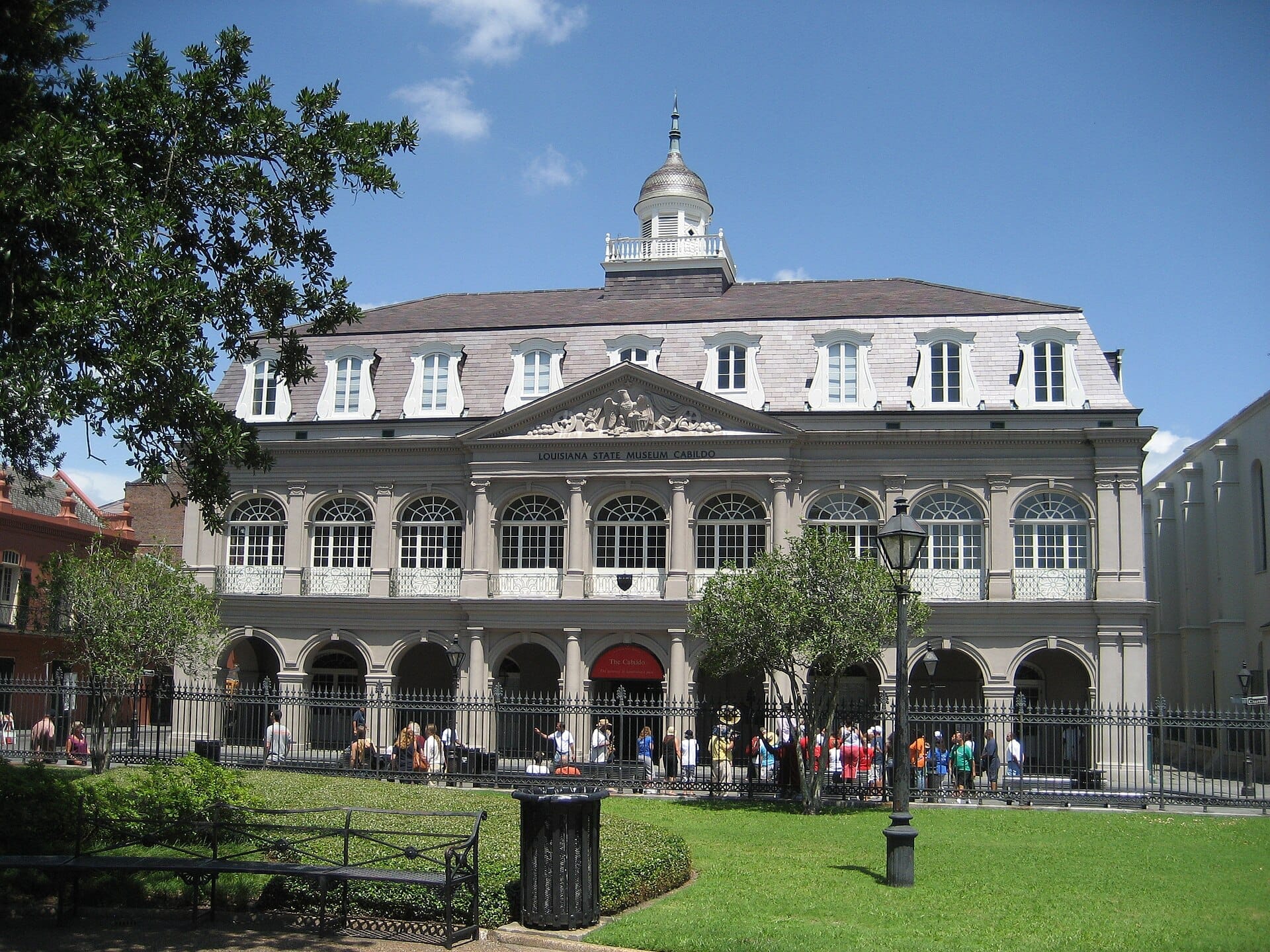 Il Cabildo a New Orleans, un grandioso edificio storico con finestre ad arco e cupola, si erge dietro una recinzione in ferro e prati ben curati in una giornata di sole.