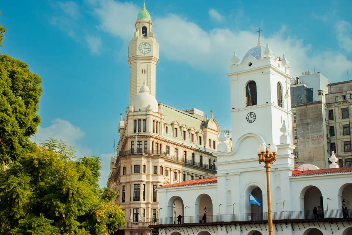 ภาพ Cabildo de Buenos Aires ยามกลางวัน อาคารสีขาวสไตล์อาณานิคม มีหน้าต่างโค้งและหอนาฬิกา ท่ามกลางท้องฟ้าสีฟ้าและตึกประวัติศาสตร์รอบข้าง