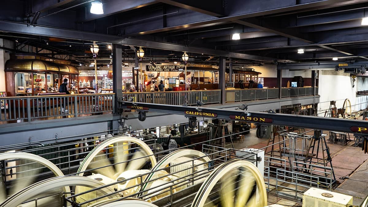 Vista interior del Museo del Teleférico de San Francisco, con ruedas de cable giratorias, maquinaria y visitantes en un balcón superior con vistas a las exposiciones antiguas.