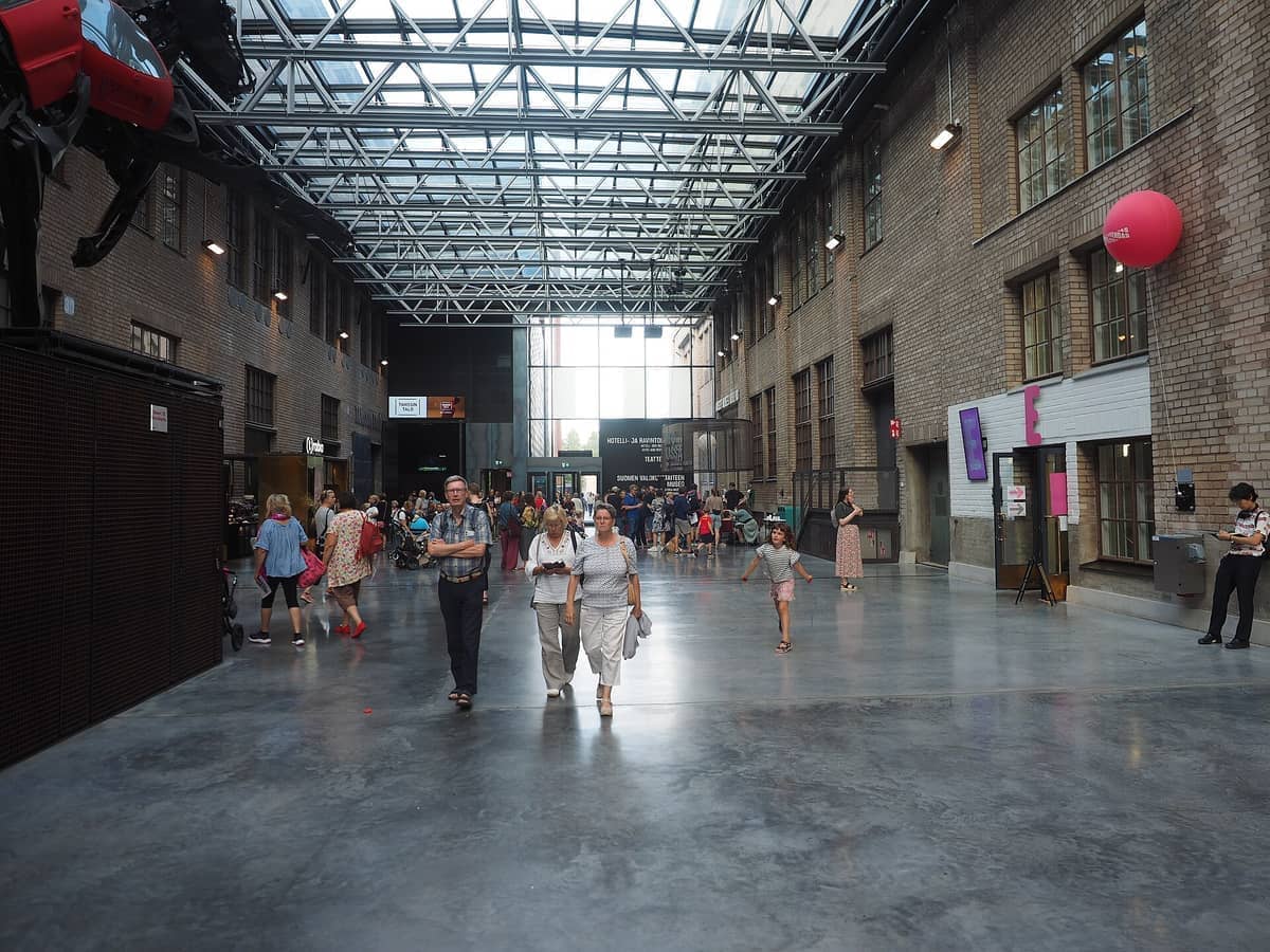 Grande salão de estilo industrial na Cable Factory em Helsinki, com paredes de tijolos aparentes, teto de vidro alto e visitantes caminhando no interior.