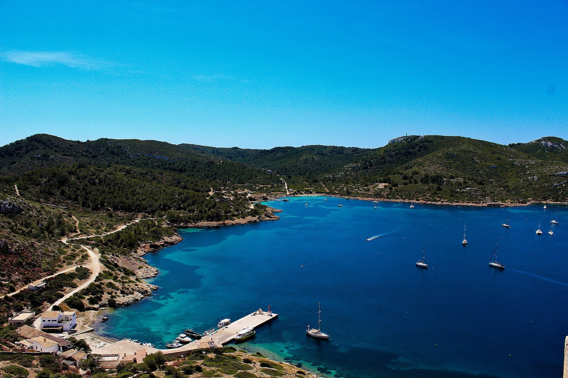 Vista aerea del Parco Nazionale di Cabrera con una baia dall'acqua azzurra cristallina, colline boscose, un piccolo porticciolo e alcune barche a vela sparse.