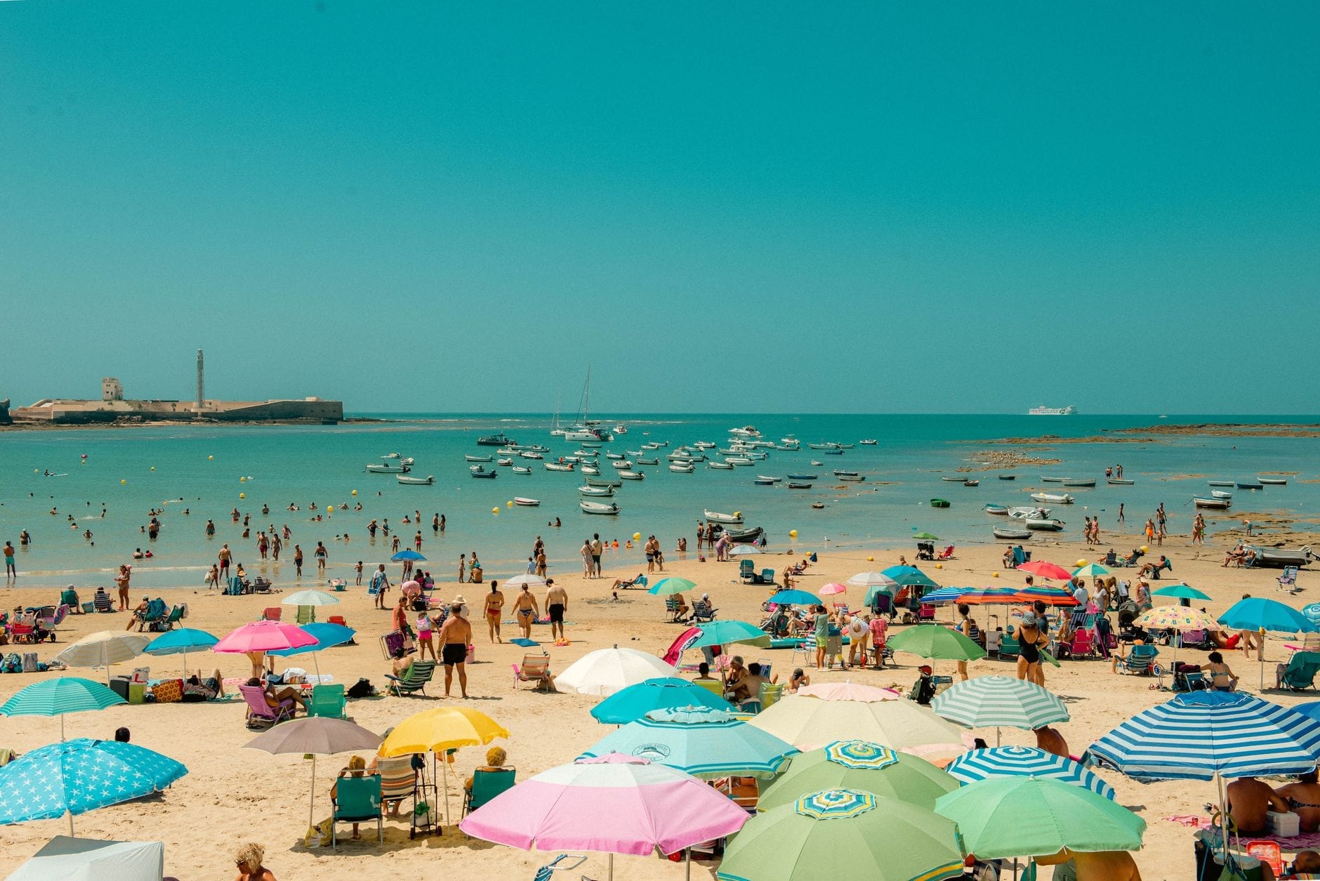 Vivace giornata estiva alla spiaggia di La Caleta a Cádiz, con ombrelloni colorati, persone che prendono il sole e nuotano, e barche ancorate al largo sotto cieli azzurri.
