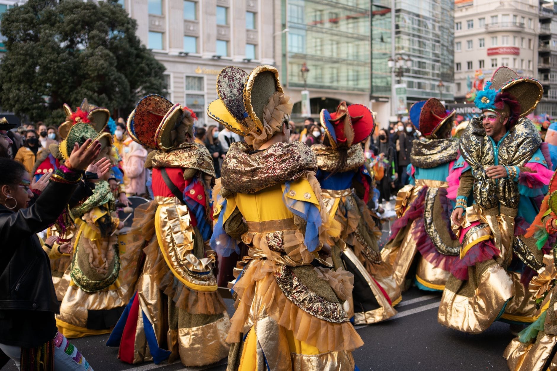 Artistas disfrazados con trajes dorados y coloridos desfilan por una calle de la ciudad con edificios al fondo y multitudes observando.