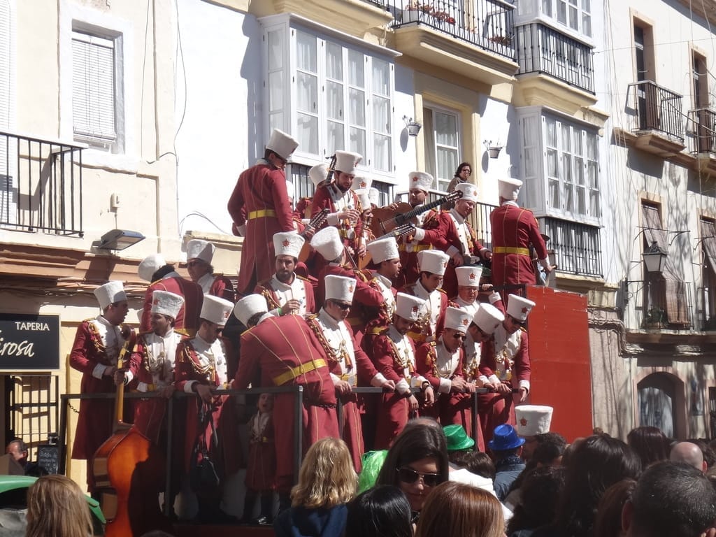 Un gruppo di musicisti in costumi coordinati rossi e bianchi si esibisce su un carro durante il Carnevale di Cádiz, con folla colorata ed edifici storici nelle vicinanze.