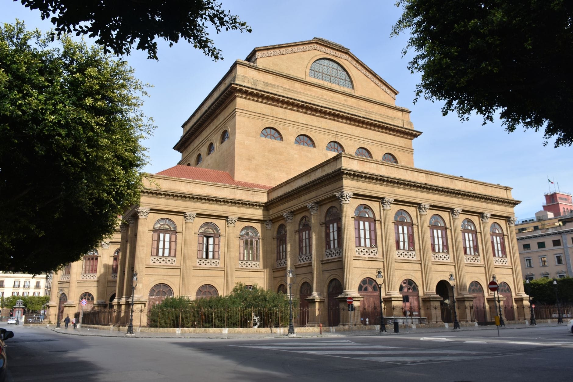 Un gran teatro histórico con ventanas arqueadas en Cádiz, España, visto desde la calle y rodeado de árboles y edificios.