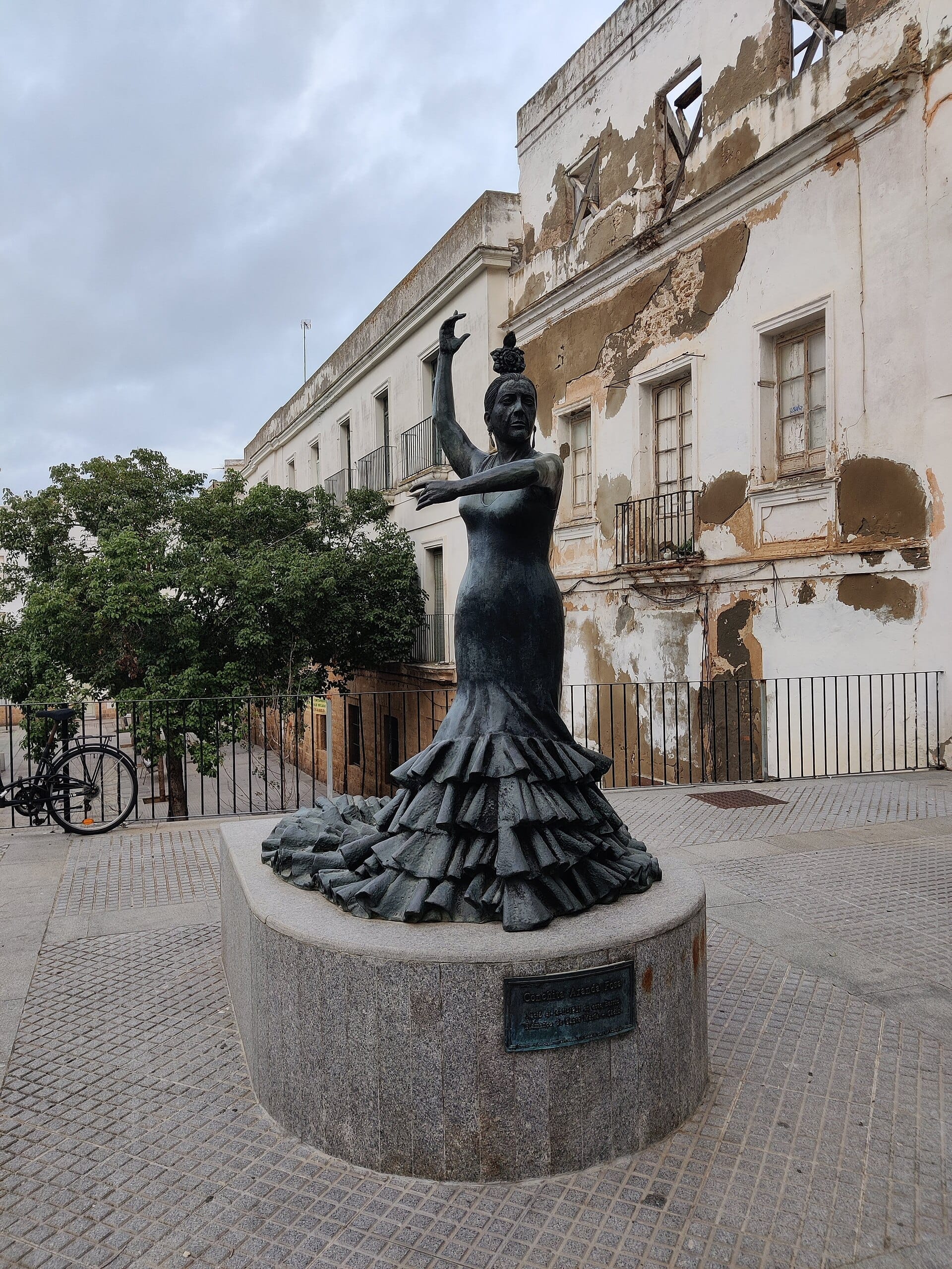 Statua in bronzo di una ballerina di flamenco a Cádiz, Spagna, con sullo sfondo un vecchio edificio deteriorato con vernice scrostata e cielo nuvoloso.