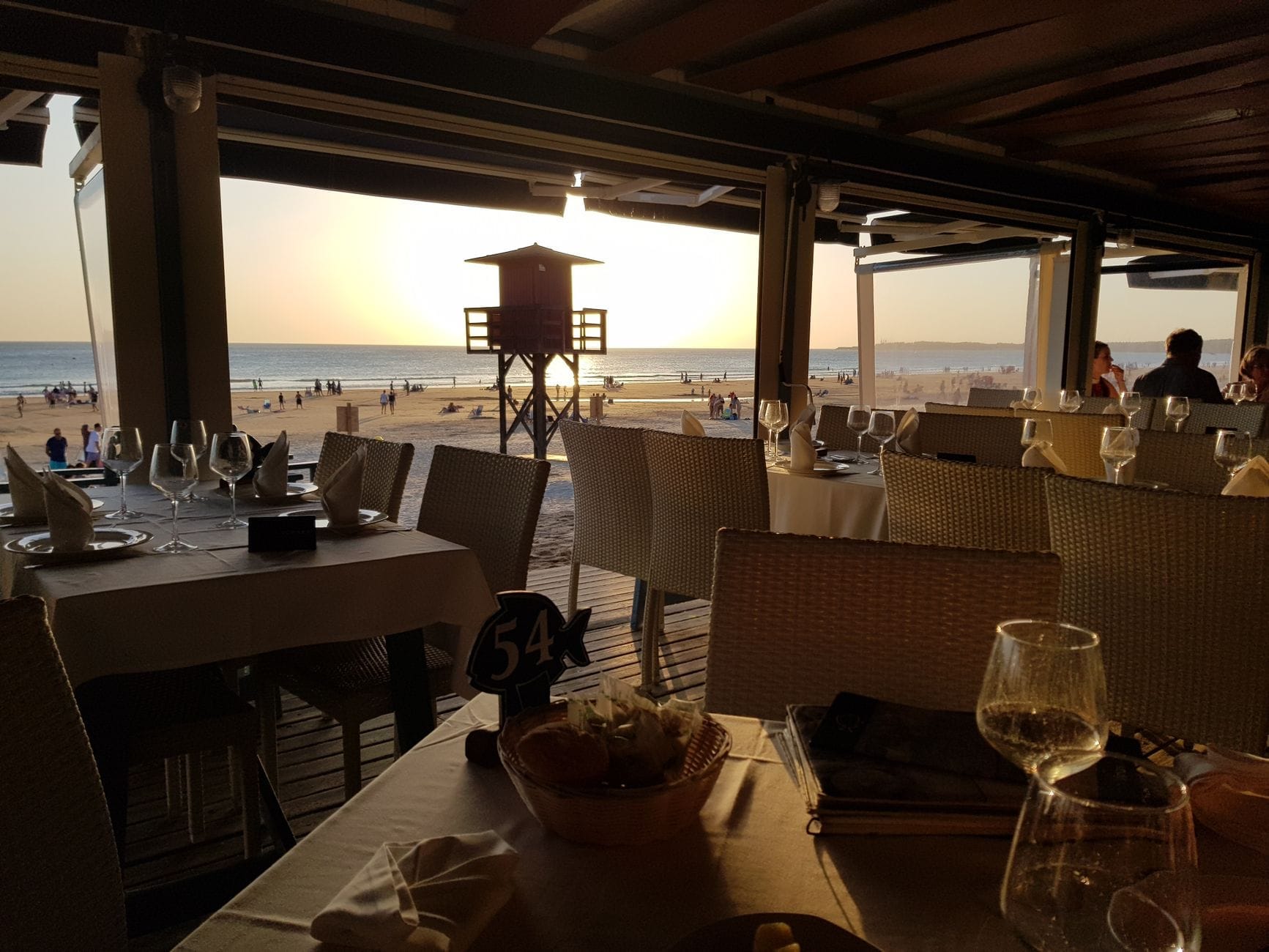Restaurante con vistas a la playa al atardecer con mesas preparadas, cristalería y personas cenando dentro y fuera