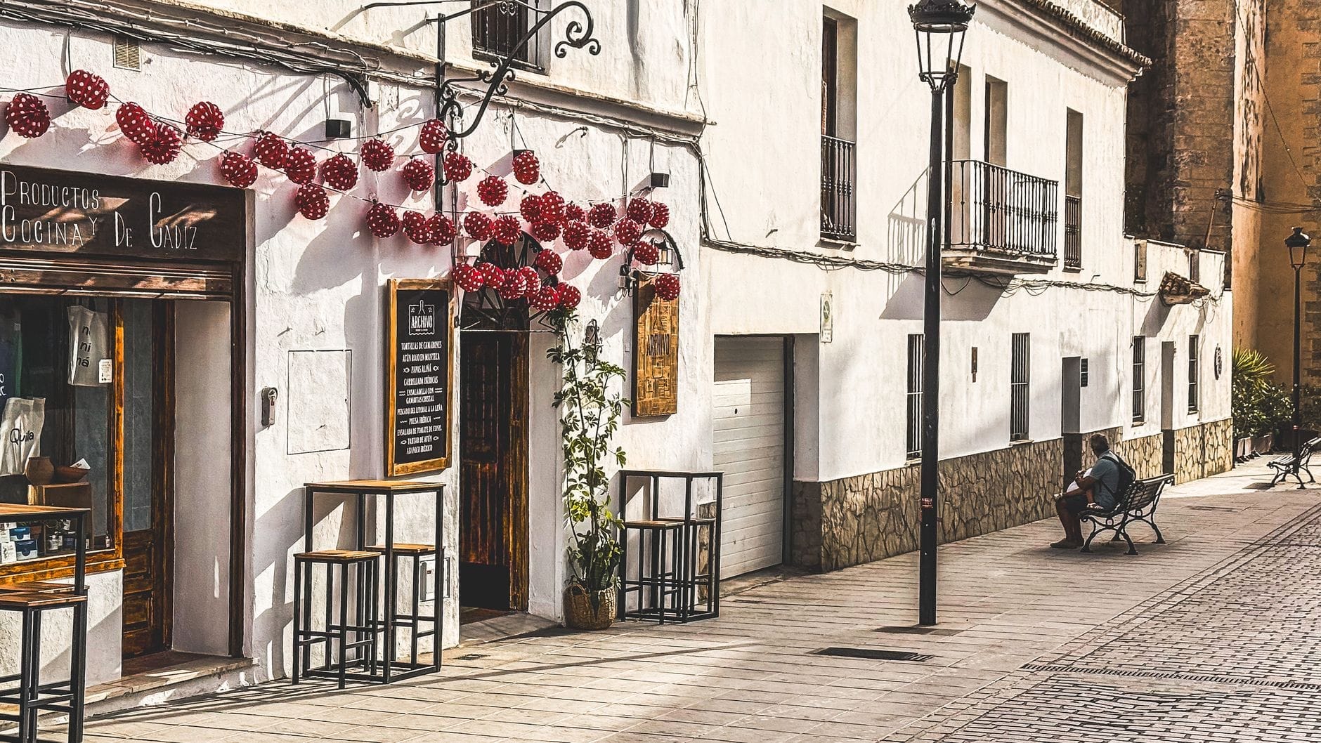 Un pequeño bar o restaurante con taburetes en el exterior, luces de guirnalda, edificios encalados y una persona sentada en un banco en una tranquila calle de Cádiz.