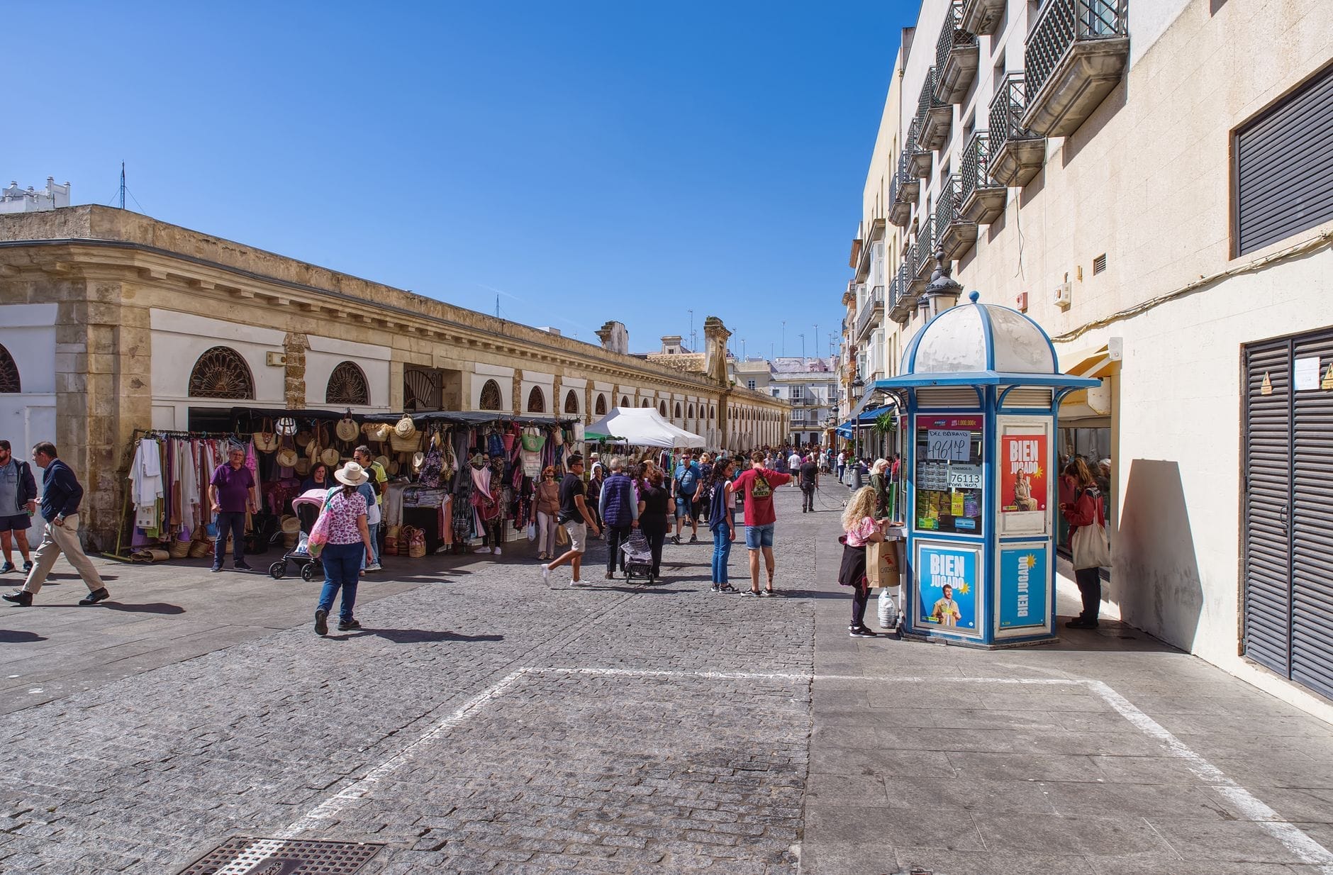 Scena di mercato all'aperto a Cádiz con persone che fanno acquisti tra bancarelle di cibo e artigianato sotto un cielo azzurro intenso, con tutta l'atmosfera locale.