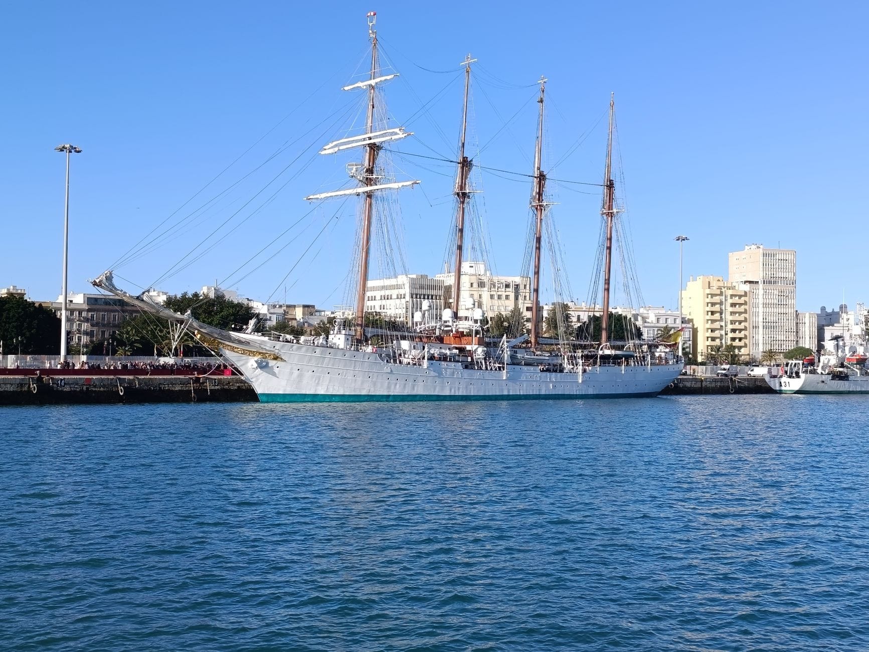 Cádiz limanında demirlemiş üç direkli klasik bir yelkenli gemi, arka planda açık mavi gökyüzü altında şehir binaları görünüyor.