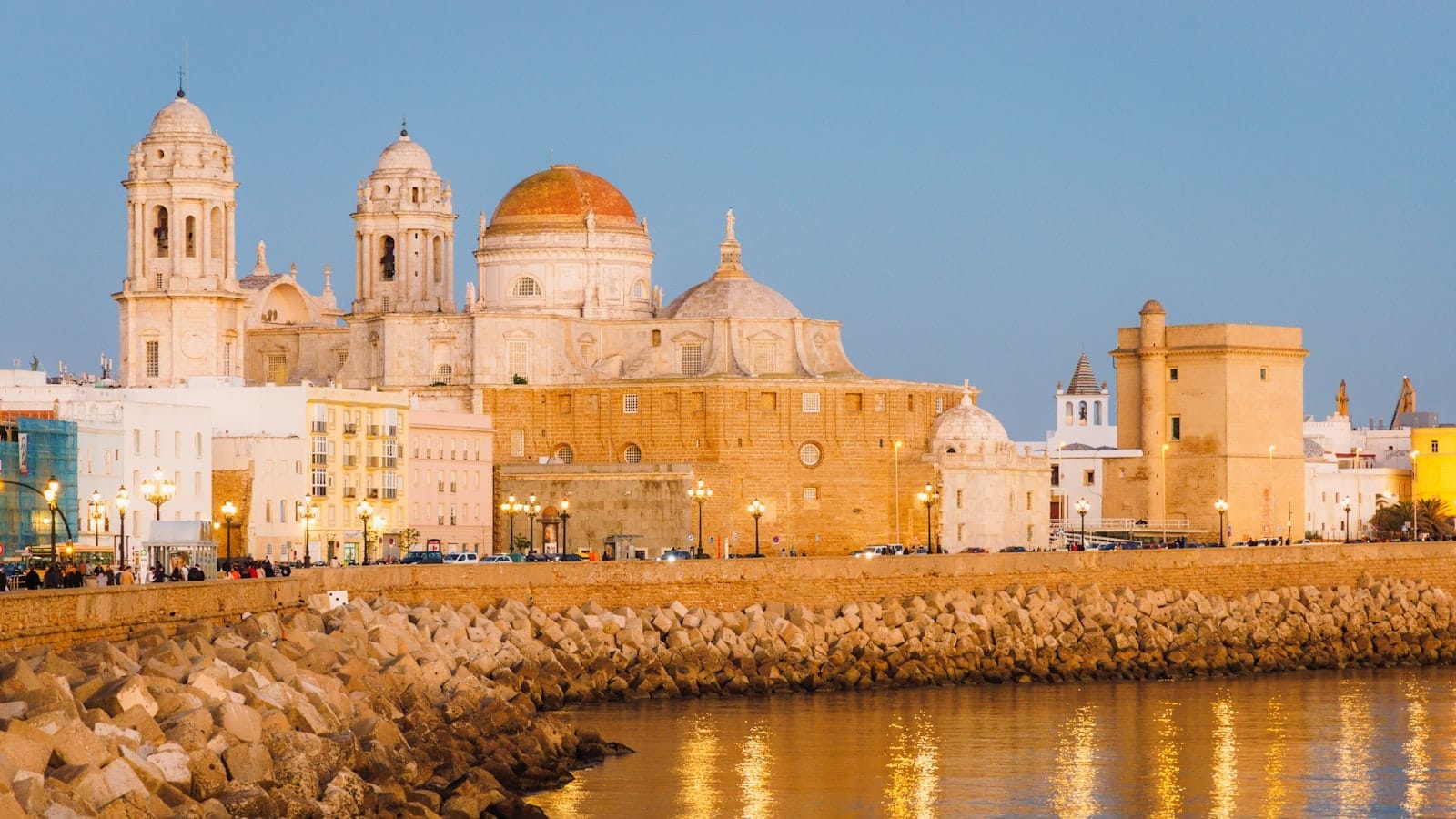 La Cattedrale di Cádiz e il suo storico lungomare illuminati dalla luce dorata della sera, visti dall'altra parte delle mura in pietra, con un cielo azzurro limpido sopra.