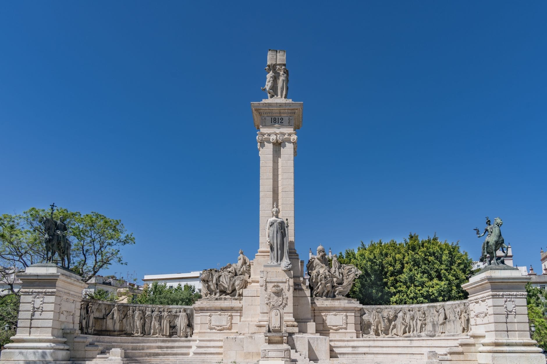 Cádiz'deki 1812 Anayasası Anıtı, açık mavi bir gökyüzünün altında heykeller ve kabartmalarla süslenmiş büyük bir beyaz taş yapı.