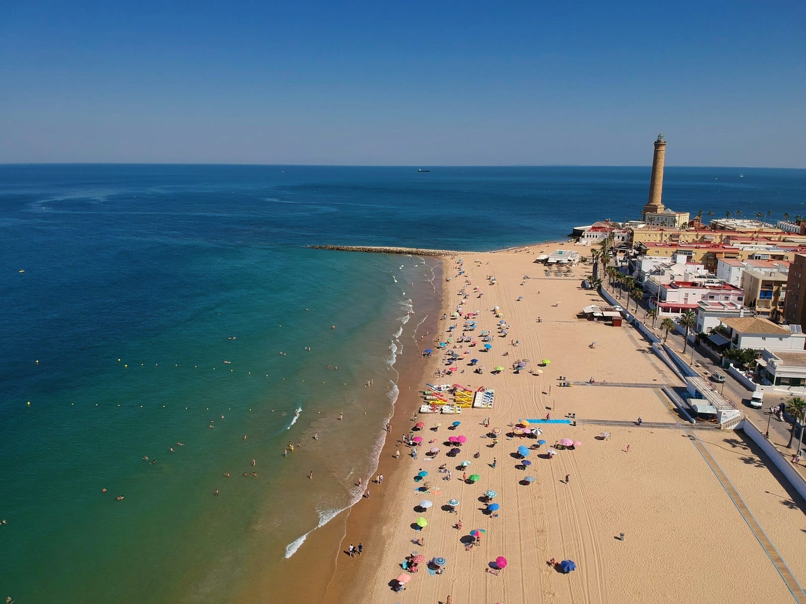 Vista aérea de una playa de arena en Cádiz con coloridas sombrillas, personas tomando el sol y bañándose, edificios de la ciudad y un alto faro al fondo.
