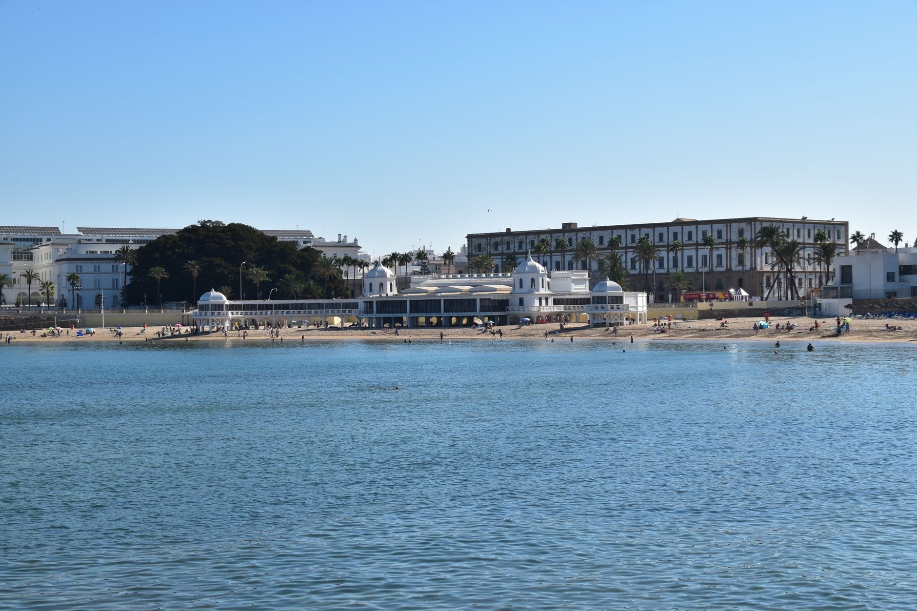 Vista de la playa La Caleta de Cádiz con el histórico pabellón de playa blanco, personas en la arena y un cielo veraniego azul y despejado.
