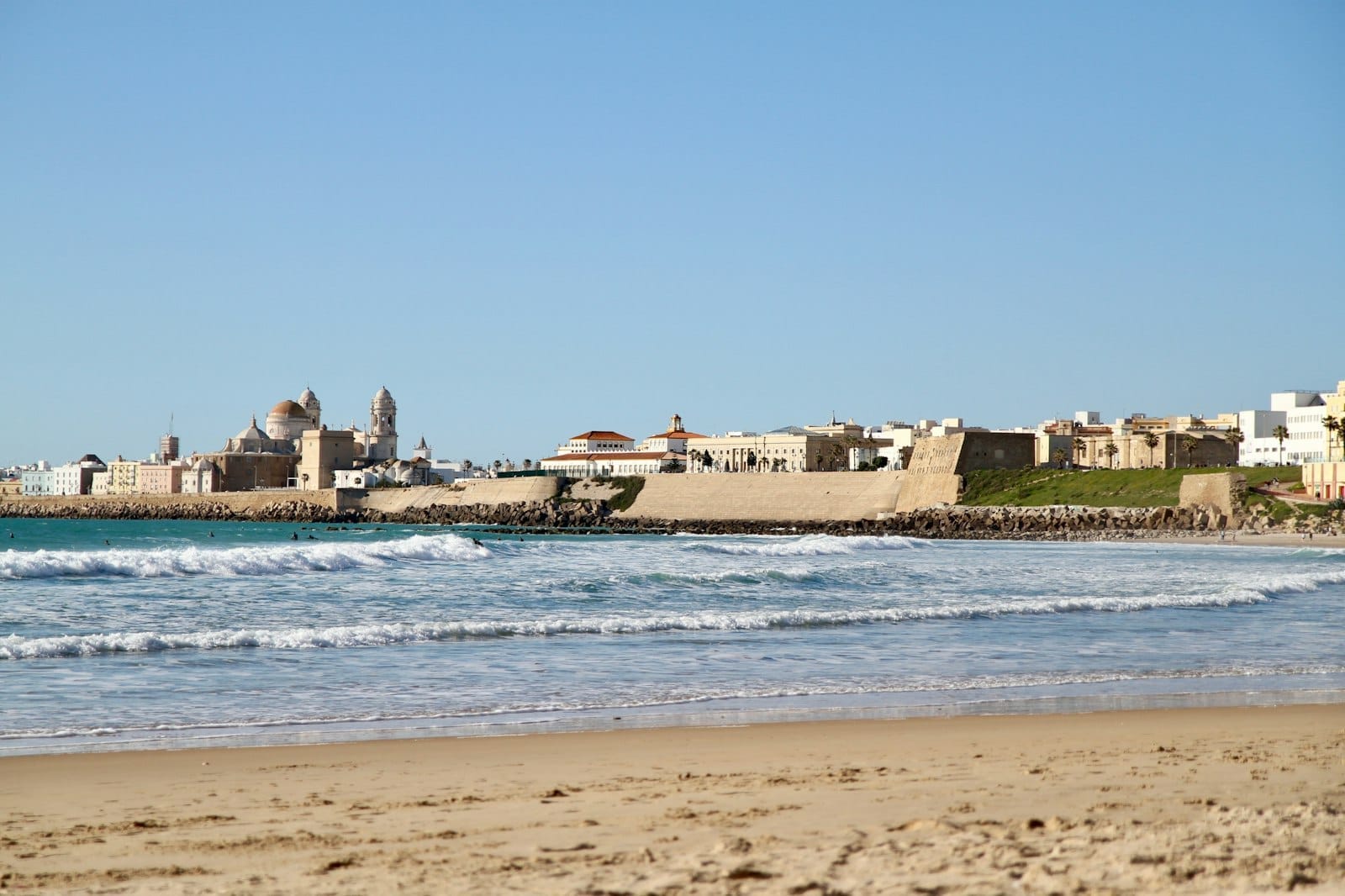 Vista panoramica della spiaggia di Cádiz con onde dolci, sabbia dorata e lo skyline storico della città sotto un cielo estivo azzurro e limpido.