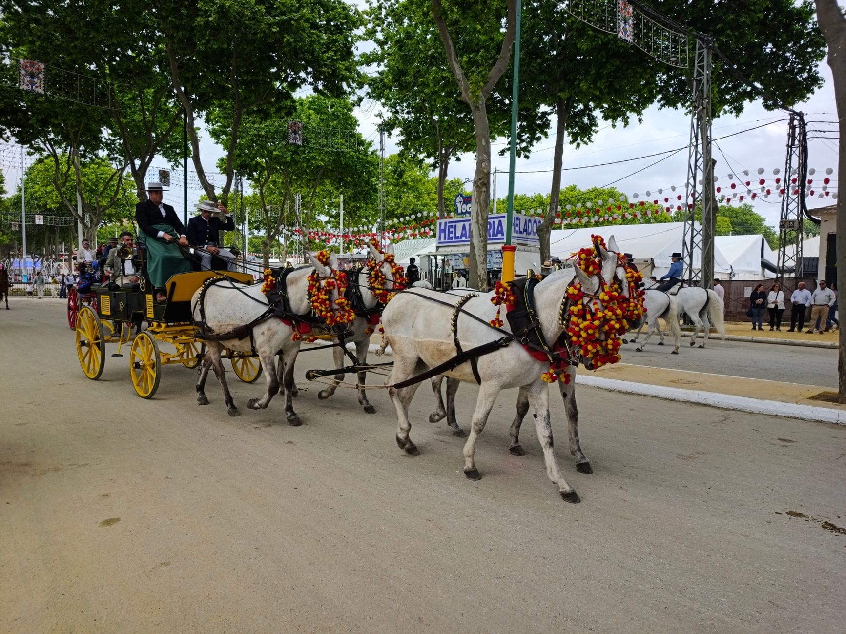 Kereta kuda yang dihiasi bunga berparade melintasi area pekan raya meriah dengan pohon, spanduk, dan orang-orang, membangkitkan suasana acara musim semi di Andalusia.
