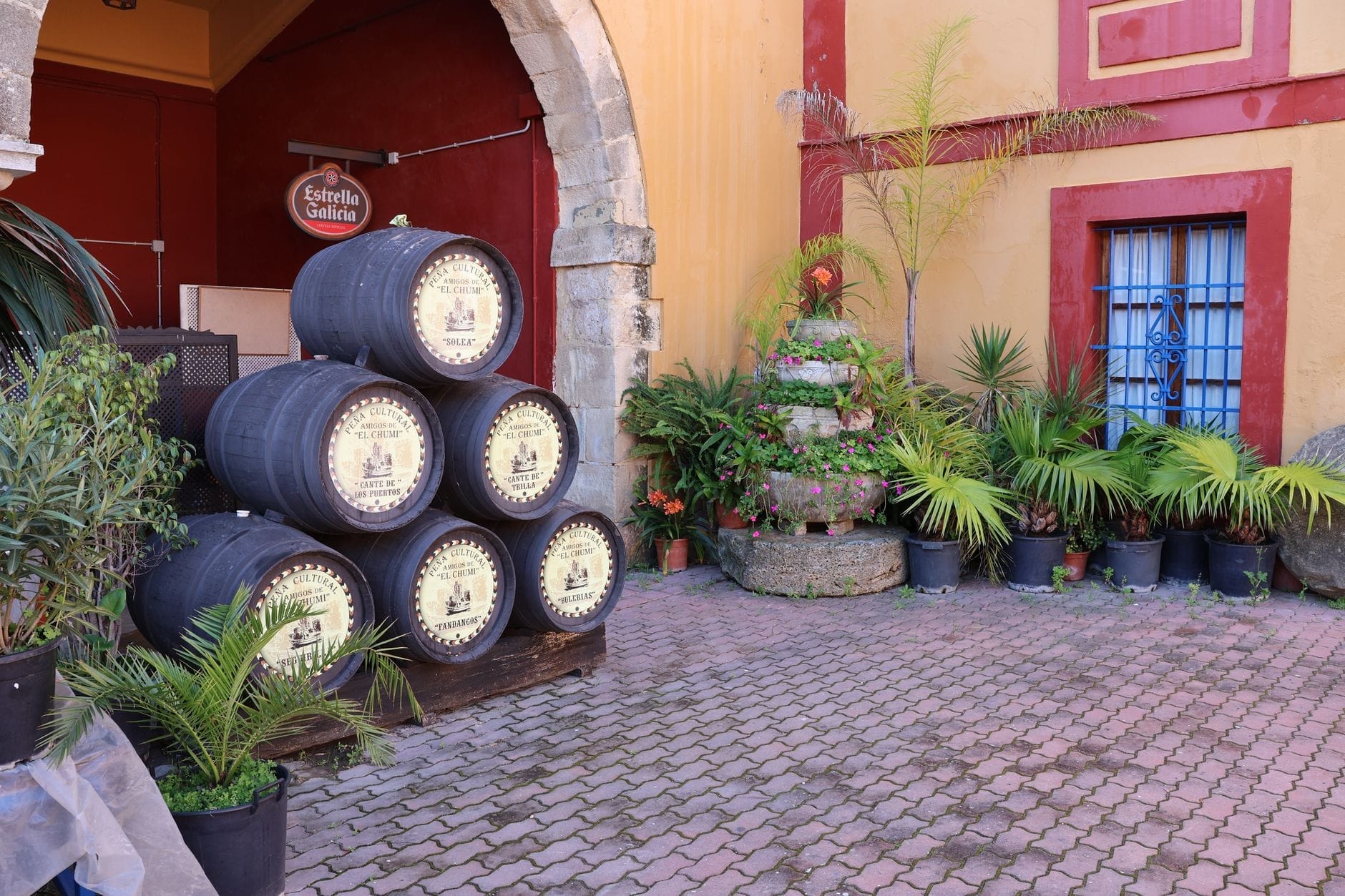 Tumpukan tong sherry di luar gedung tradisional di Jerez, dikelilingi tanaman pot dan fasad berwarna-warni.