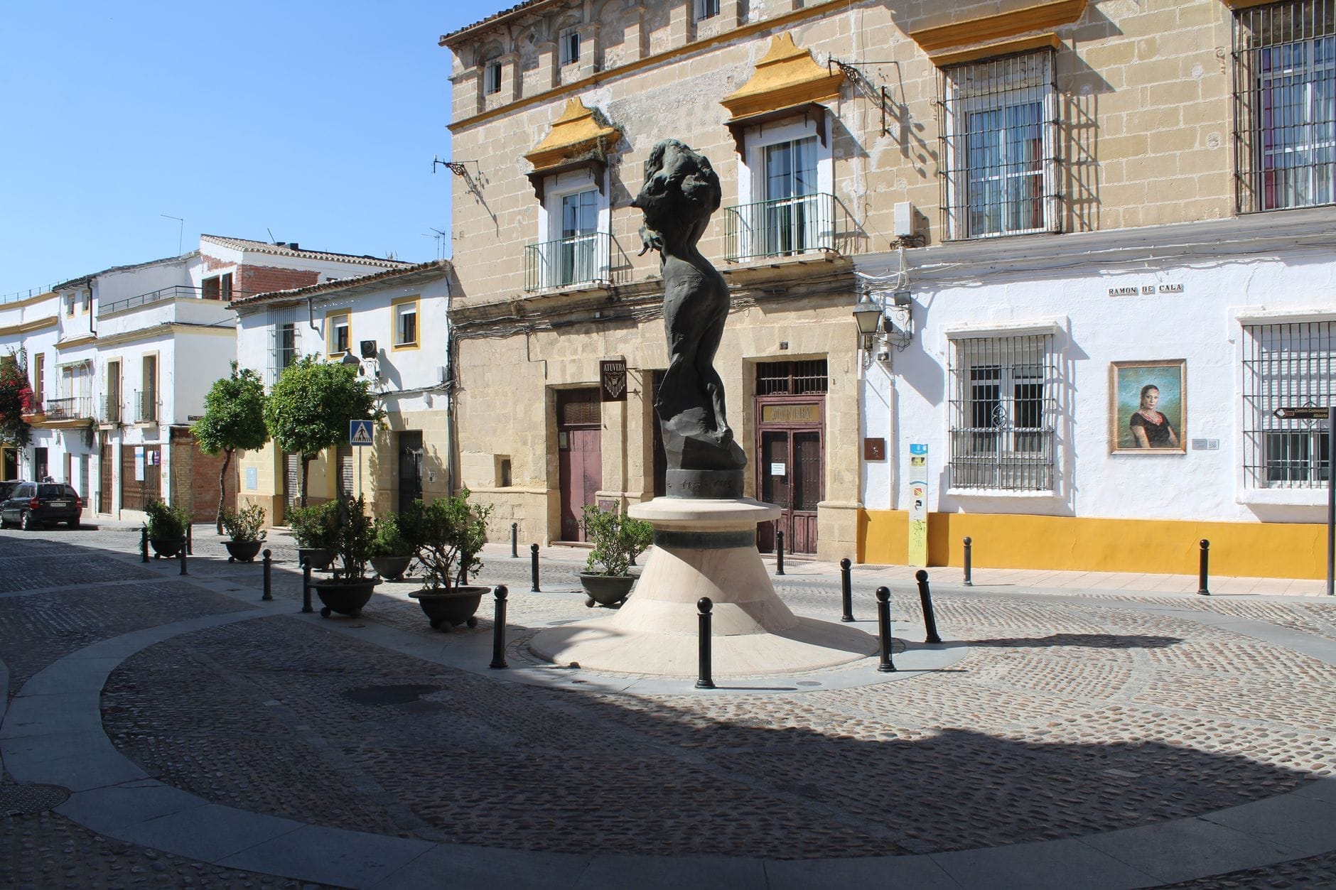 Sebuah plaza cerah di Jerez de la Frontera dengan patung modern, bangunan bergaya Andalusia, dan jalan berbatu, mencerminkan suasana kota.