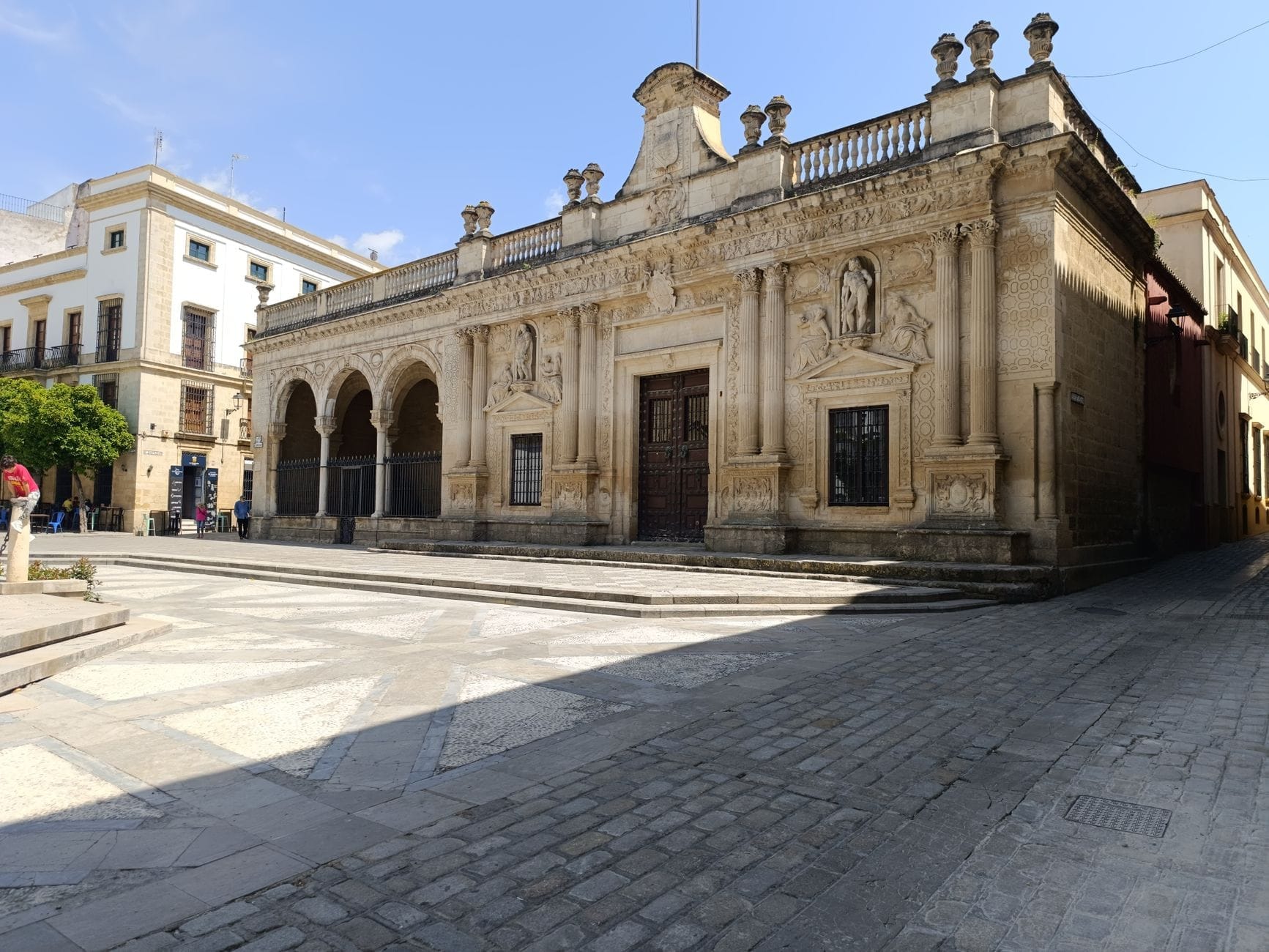 Ampia piazza con un imponente edificio storico in pietra e colonnato ad archi a Jerez de la Frontera, sotto un cielo blu, che invita all'esplorazione.