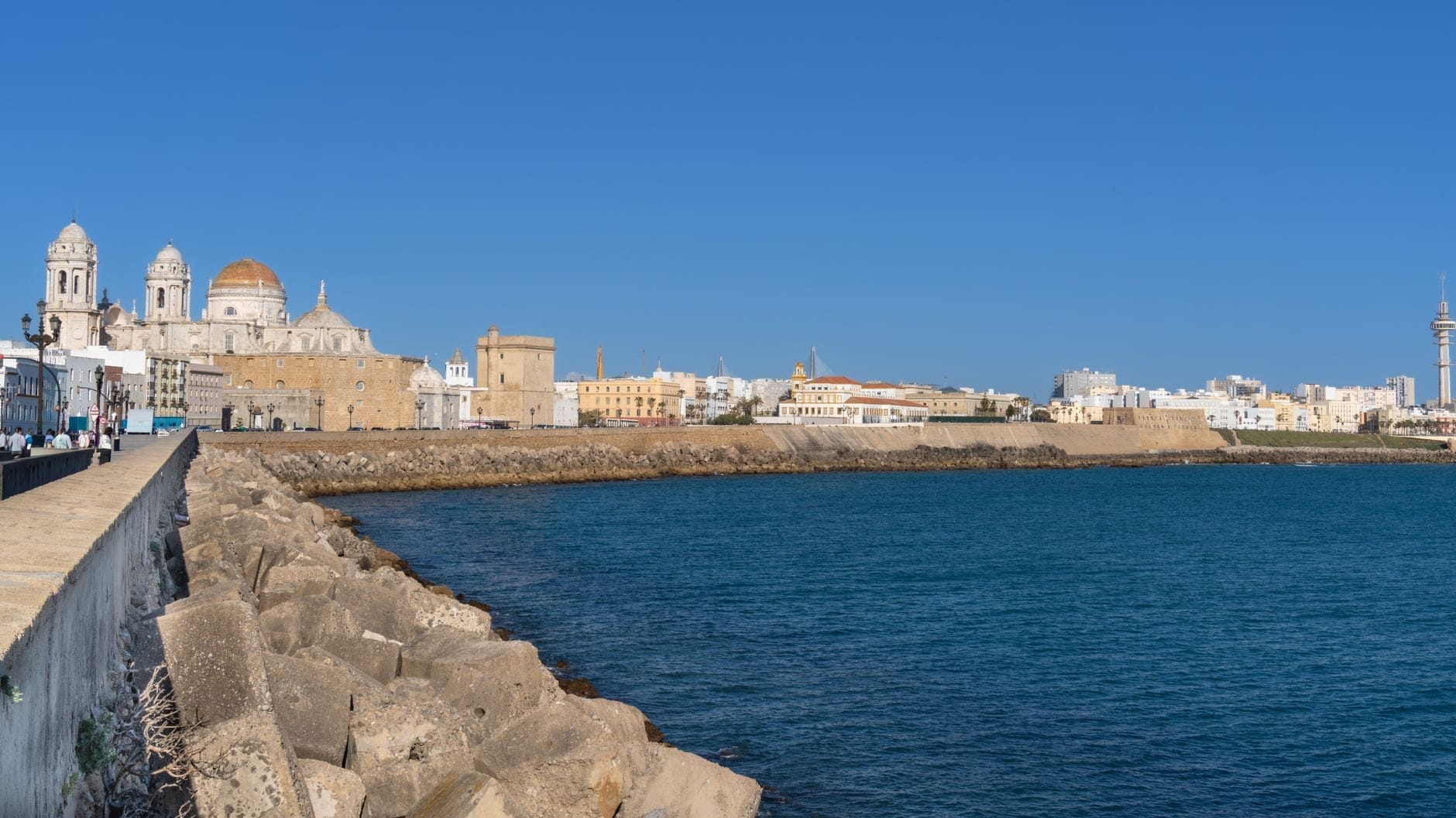 Vista panoramica del lungomare del centro storico di Cádiz con le cupole della cattedrale, gli edifici storici e il paseo marittimo sotto un cielo azzurro limpido.