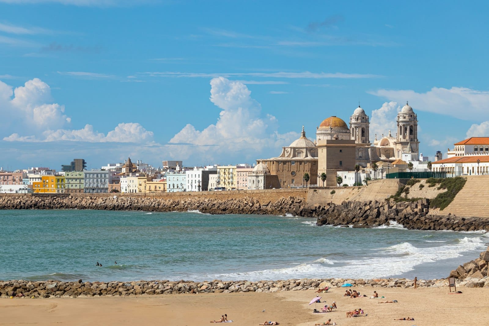 Panoramic view of Cadiz with the iconic cathedral, colorful waterfront buildings, sandy beach, and blue sky, capturing the essence of the Spanish coastal city.