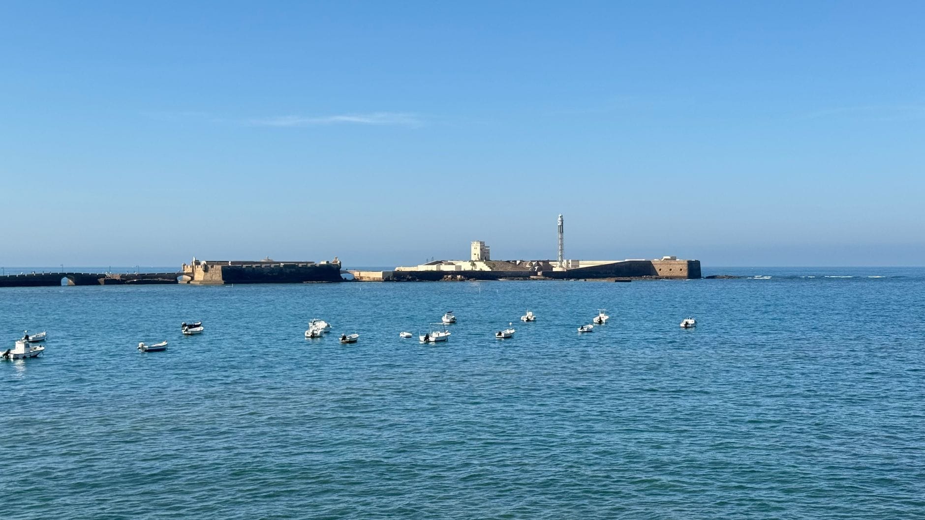 View of a historic seaside fortress with boats anchored in calm blue waters under a clear sky, Cádiz, Spain.