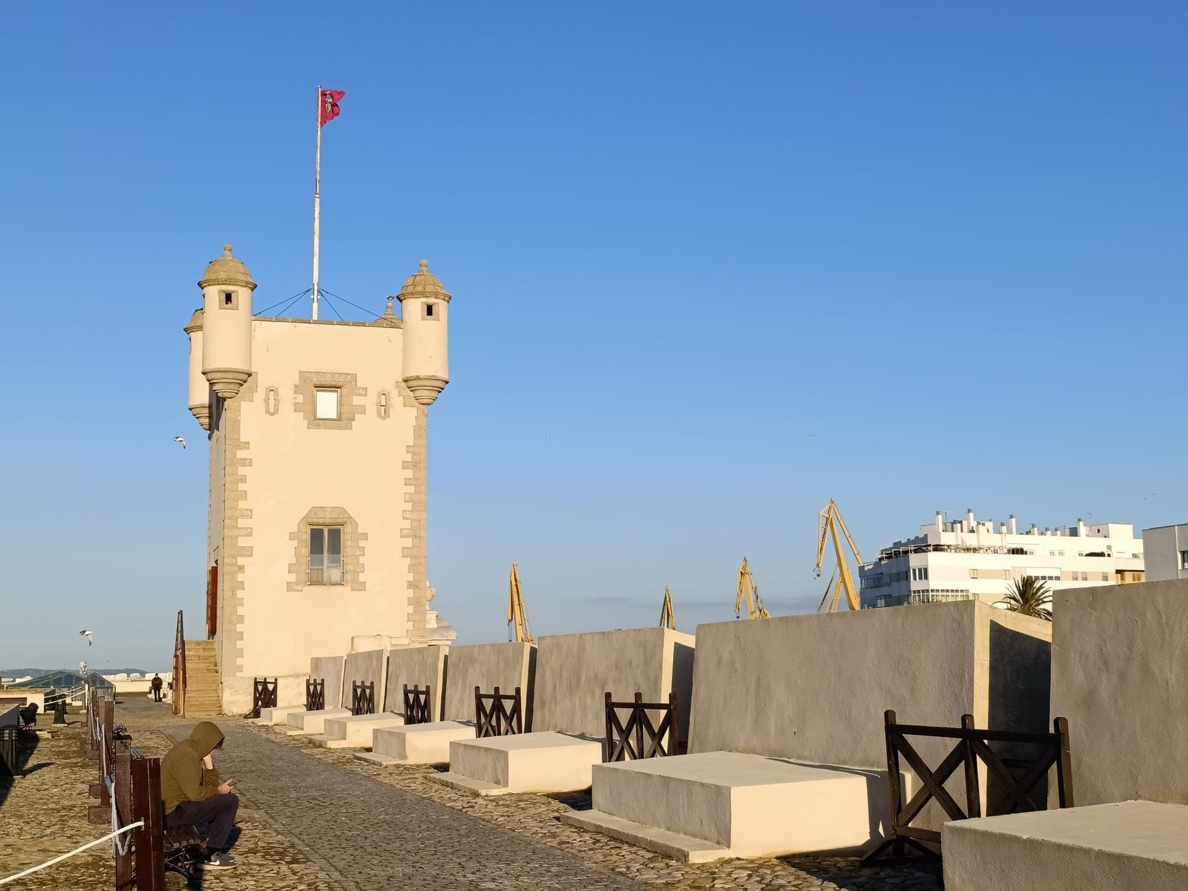 Historic watchtower with two turrets and a flag, facing benches and a coastal promenade under a clear blue sky in Cádiz, Spain.