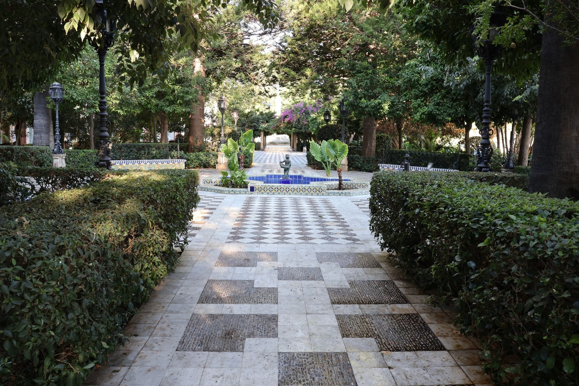 Quiet park walkway lined with hedges, patterned pavement and central fountain, shaded by trees in Cadiz, Spain.
