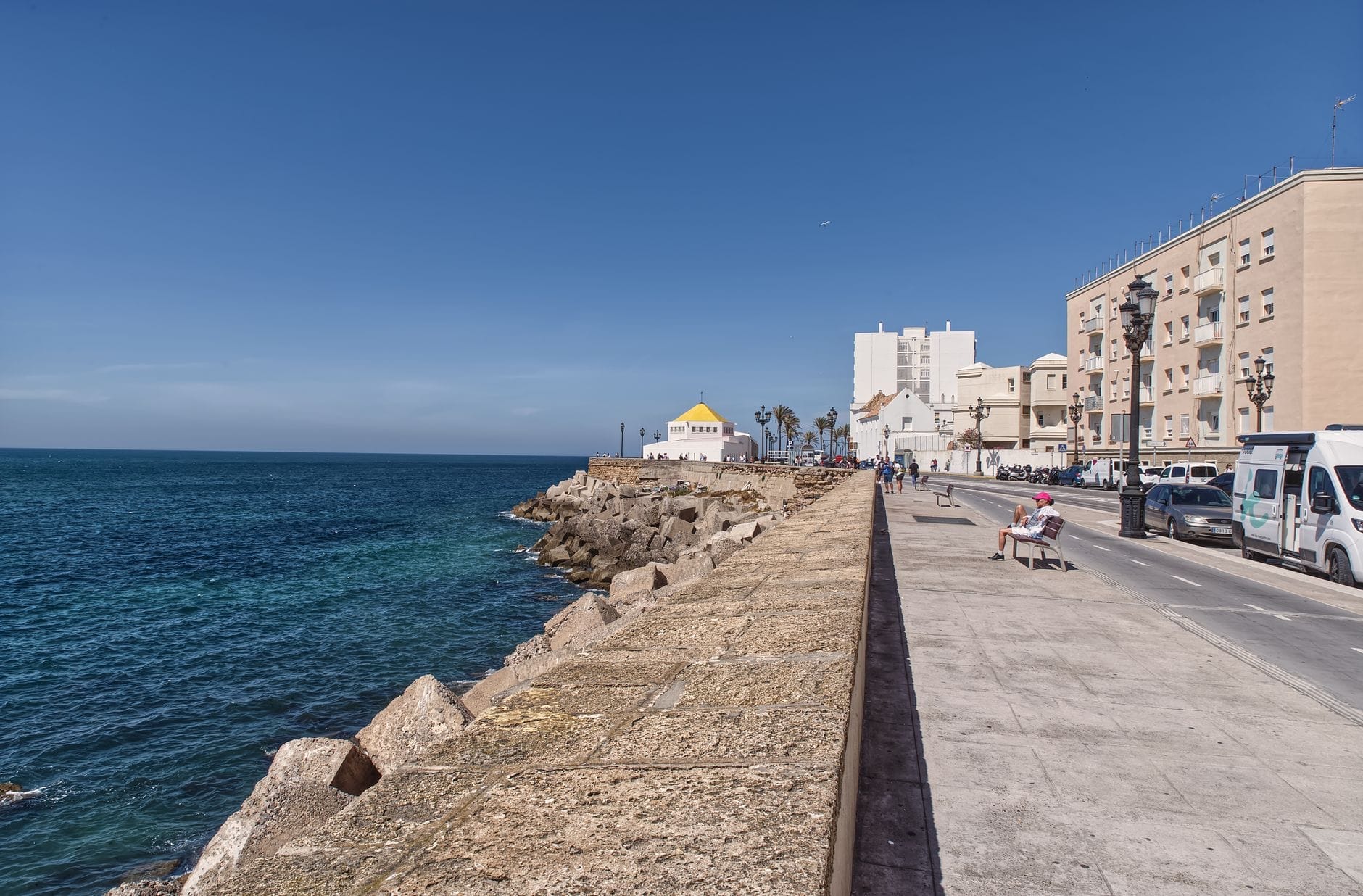 Seaside promenade in Cádiz with stone sea wall, rocky shore, and people walking beside the ocean, city buildings in background under clear blue sky.