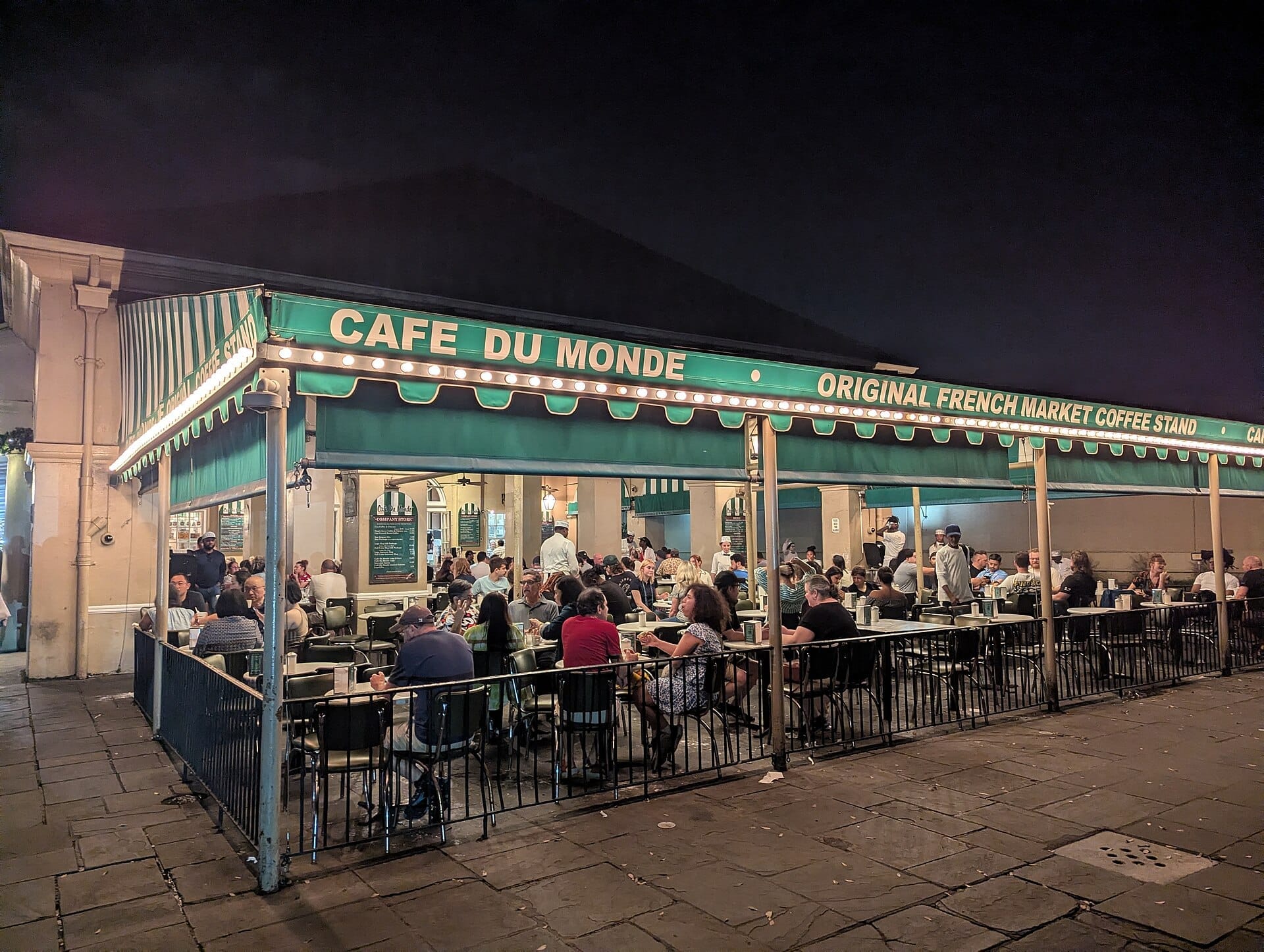 L'area dei posti a sedere all'aperto del Café du Monde di notte, con persone che gustano beignets e caffè sotto la tenda verde e le luci d'atmosfera.