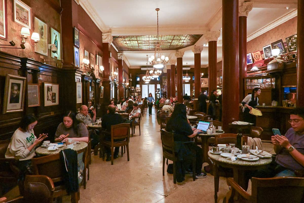 บรรยากาศภายใน Café Tortoni ที่บัวโนสไอเรส มีผู้คนจำนวนมากนั่งตามโต๊ะใต้โคมไฟระย้าและเพดานกระจกหลากสี ตกแต่งด้วยไม้เข้มและภาพศิลปะกรอบใหญ่