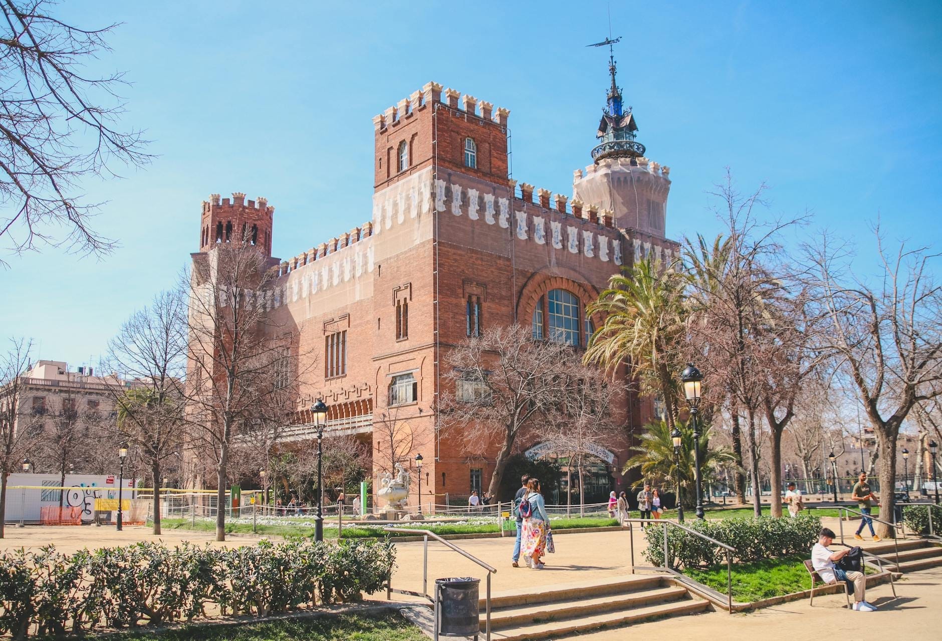 Distinctive red brick modernista building with turrets and ornamental details, surrounded by trees and people enjoying a sunny day in Barcelona.
