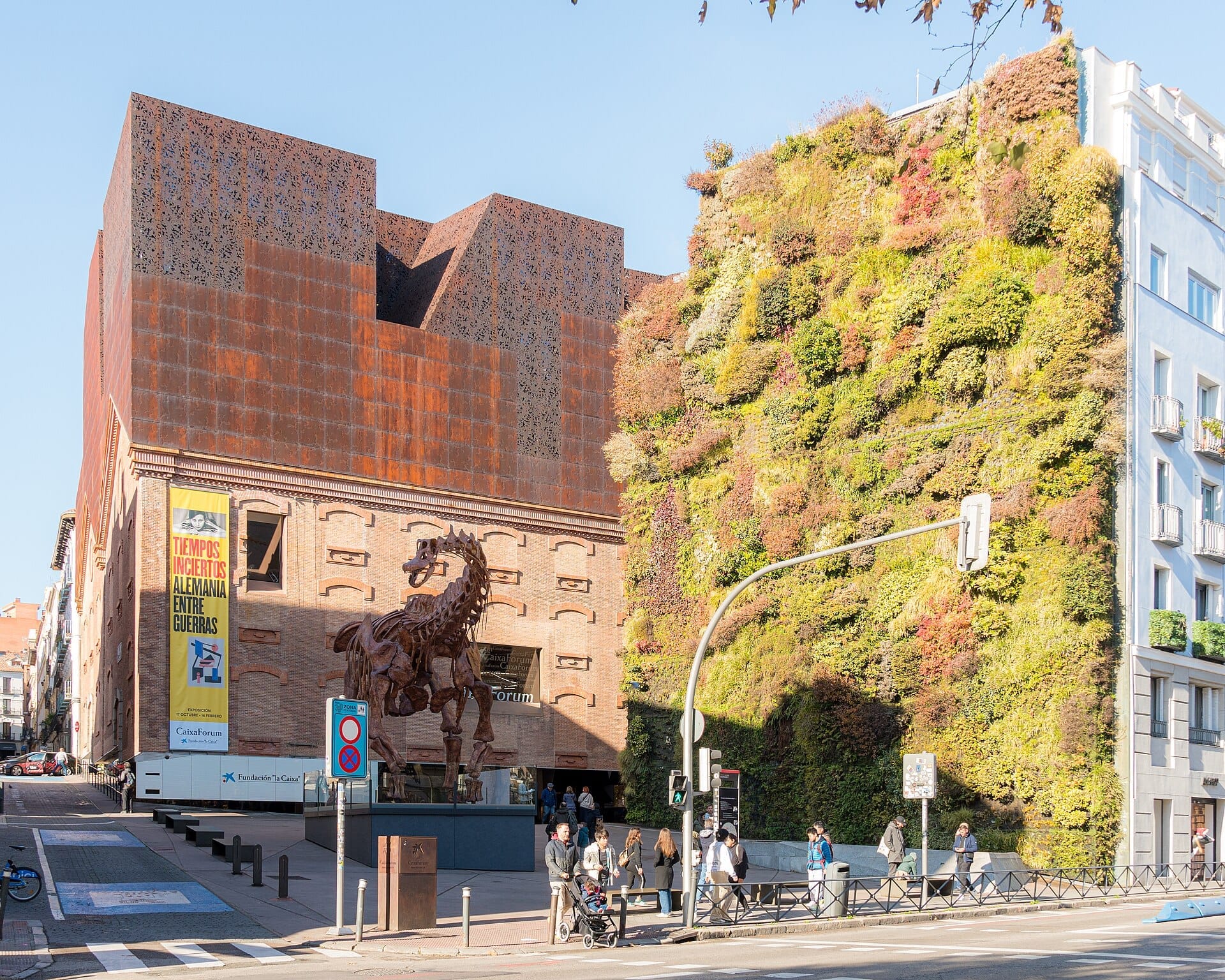 Esterno del CaixaForum Madrid con la sua caratteristica architettura color ruggine, un grande giardino verticale, persone a passeggio e una prominente scultura di un cavallo sotto un cielo azzurro limpido.