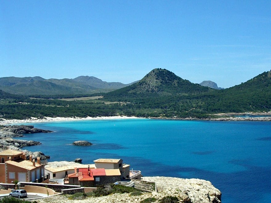 Vista della spiaggia Cala Agulla a Mallorca con acqua turchese, sfondo di pineta, colline e alcune case lungo la riva rocciosa.