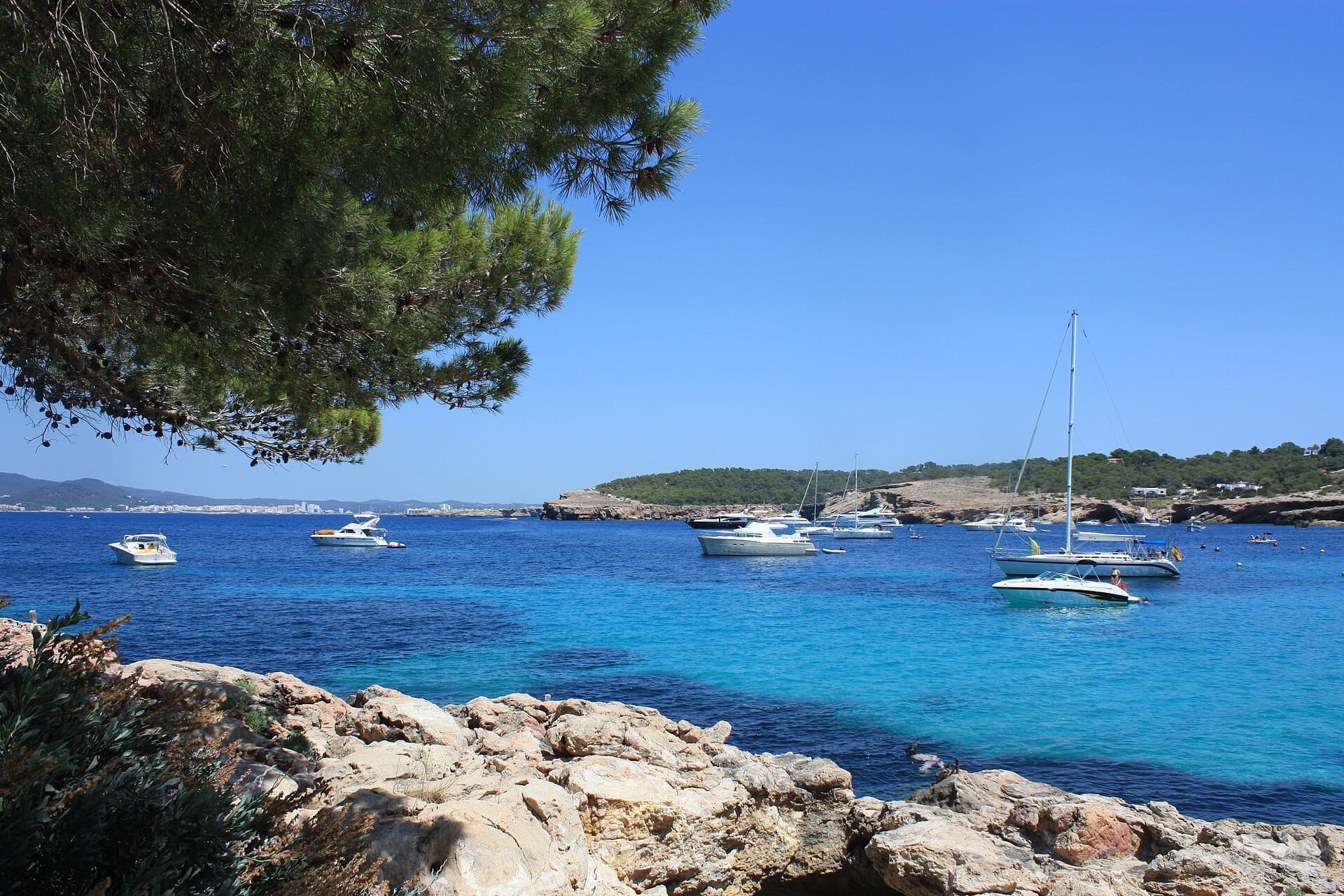 Acqua azzurra cristallina e barche ancorate viste dalla riva rocciosa con pini alla Cala Bassa Beach, Ibiza, sotto un cielo luminoso e soleggiato.