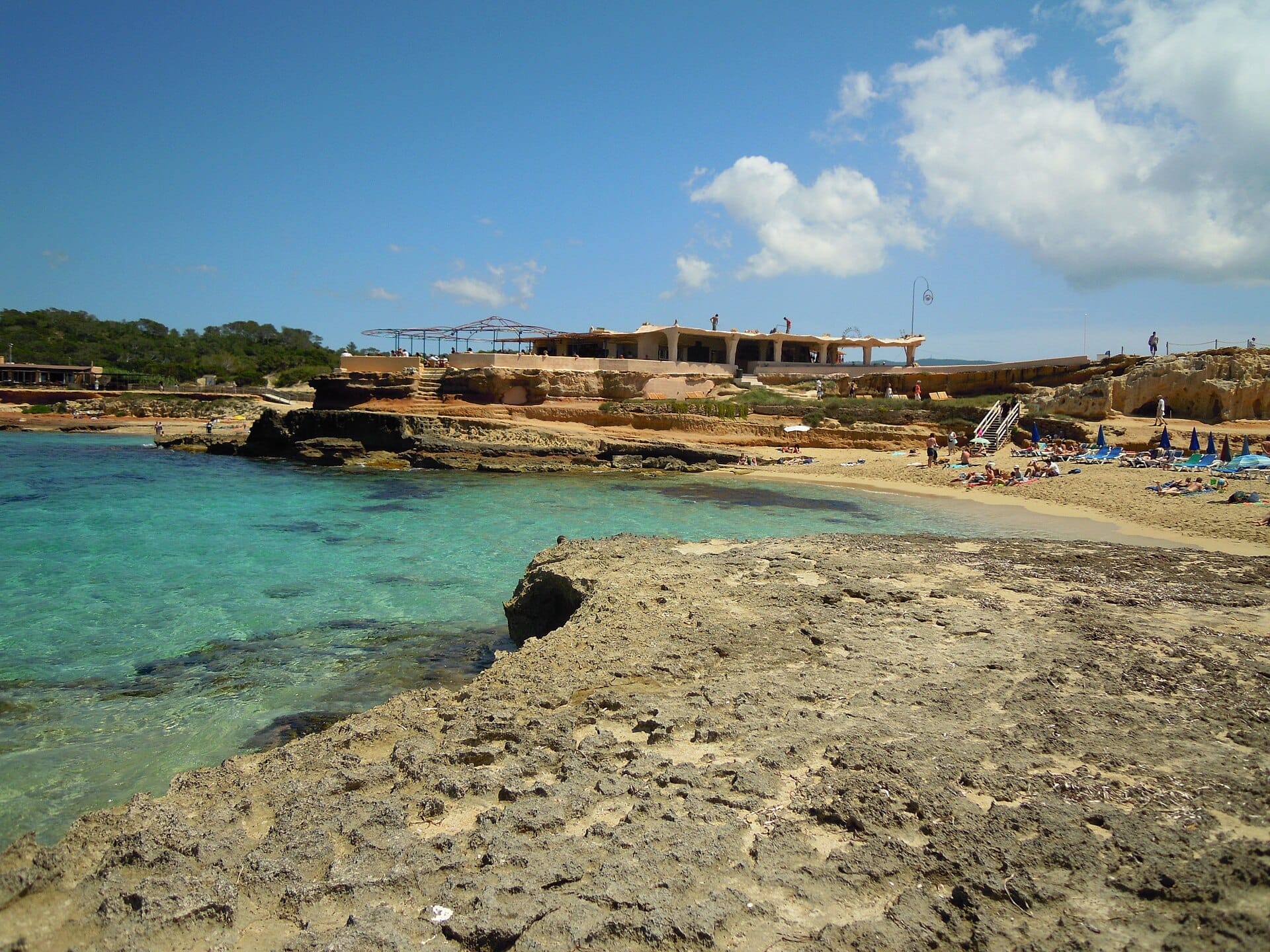 Vista della spiaggia di Cala Comte a Ibiza con acque turchesi limpide, riva rocciosa, calette sabbiose, bagnanti e un rustico ristorante sul mare.