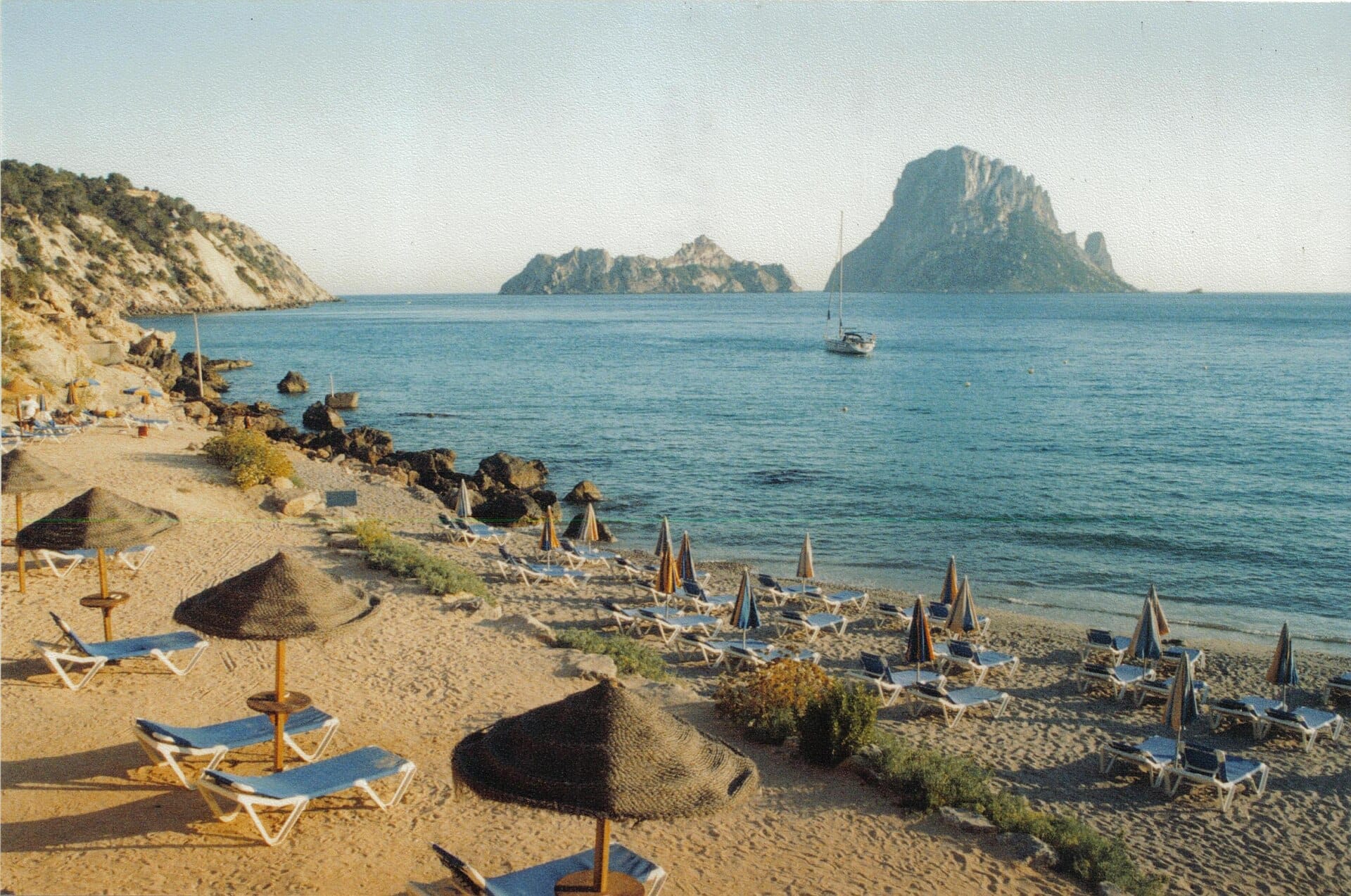 La spiaggia di Cala d'Hort con file di lettini vuoti e ombrelloni di paglia rivolti verso il mare blu e la rocciosa isola Es Vedrà sullo sfondo.