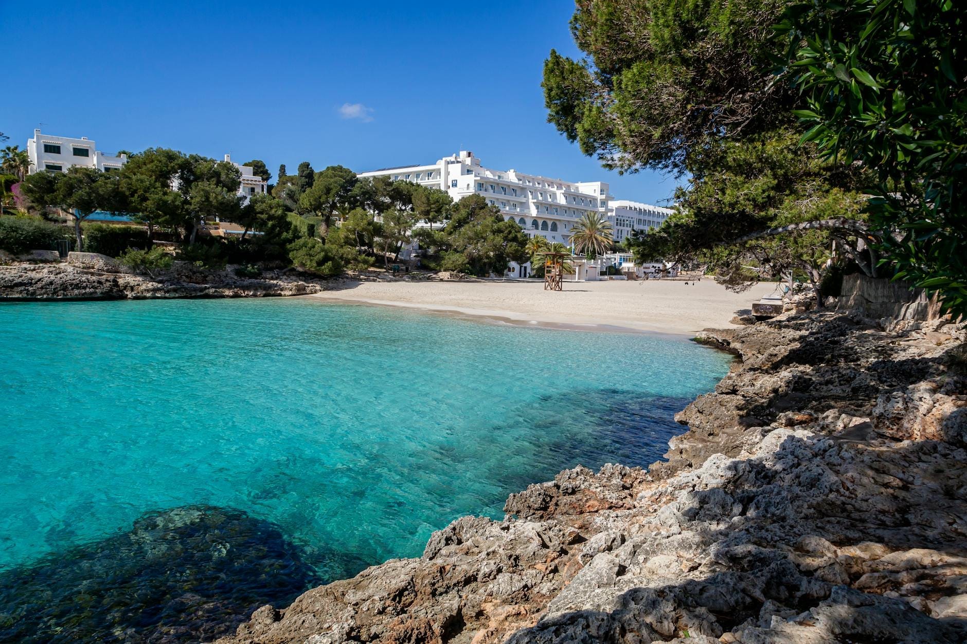 Caletta con acqua turchese, spiaggia di sabbia bianca, riva rocciosa in primo piano, edifici imbiancati a calce e alberi sullo sfondo sotto un cielo azzurro limpido.