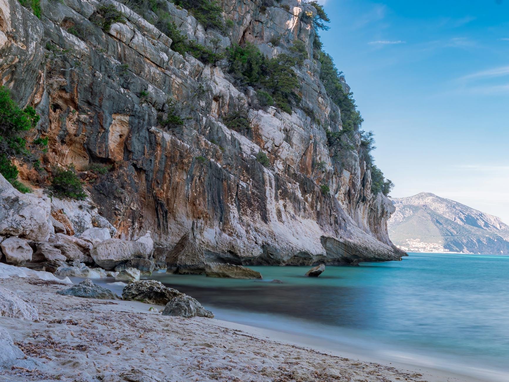 Blick auf den Kiesstrand der Cala Goloritzè mit türkisblauem Wasser, imposanten Kalksteinfelsen ringsum und einer fernen Bergküste unter strahlend blauem Himmel.