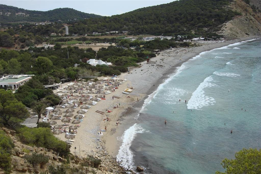 Vista aerea della spiaggia di Cala Jondal con ciottoli bianchi, acqua turchese, lettini e beach club circondati da colline verdi.