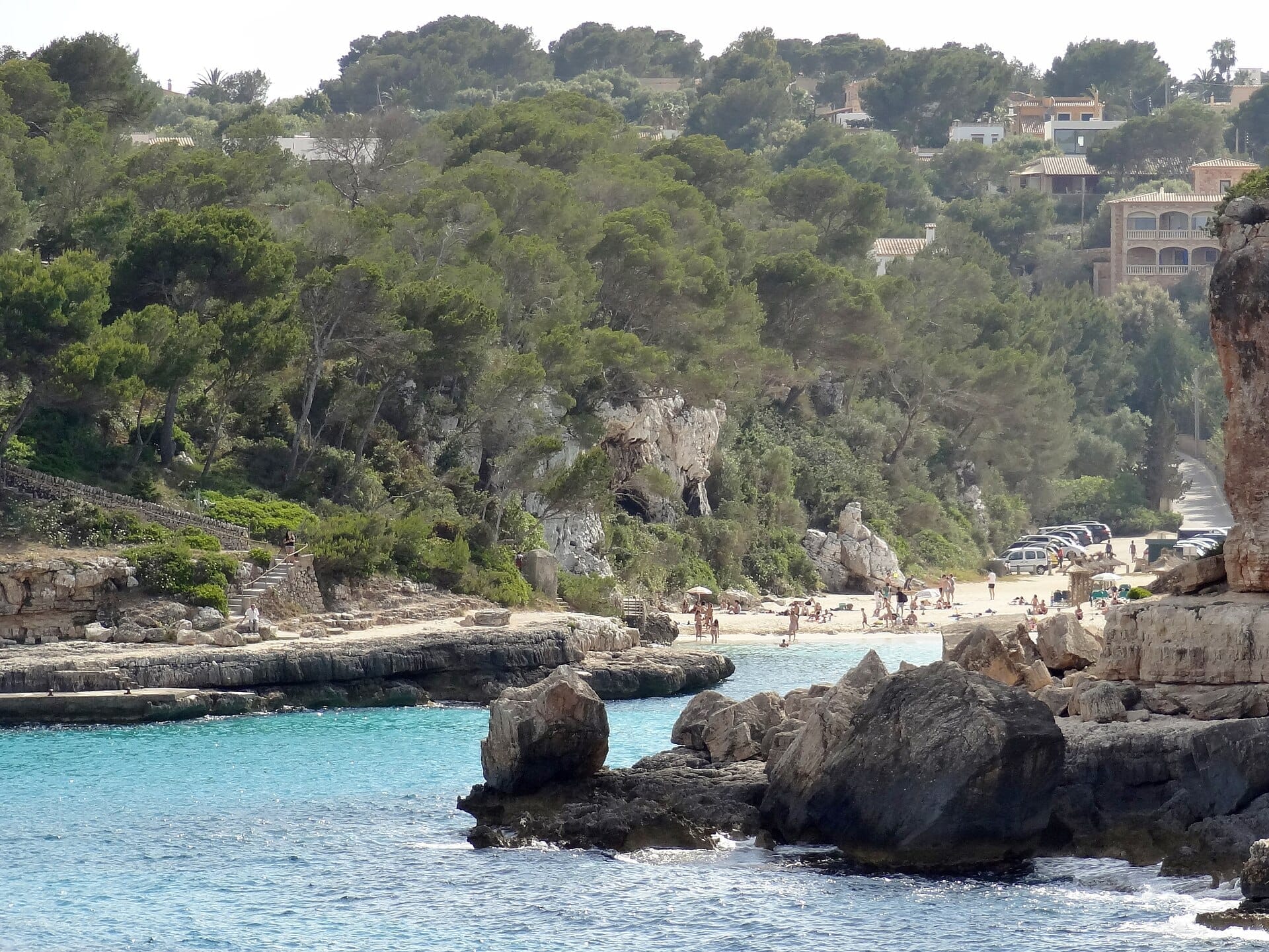 Spiaggia di Cala Llombards con acqua turchese, sabbia bianca, scogliere calcaree, bagnanti sparsi, pini e ville sul pendio sullo sfondo.