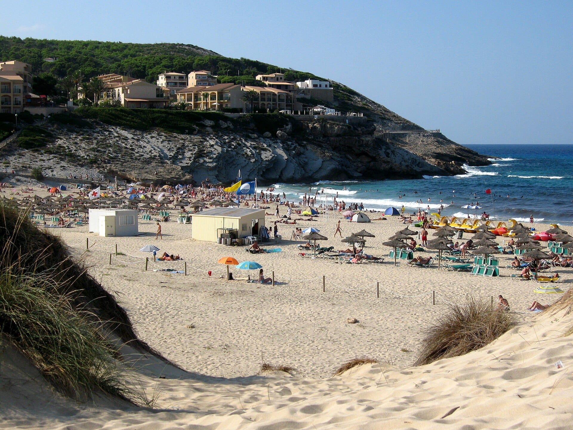 Spiaggia di Cala Mesquida con dune di sabbia dorata, lettini, ombrelloni e onde turchesi, circondata da scogliere rocciose ed edifici sul fianco della collina.