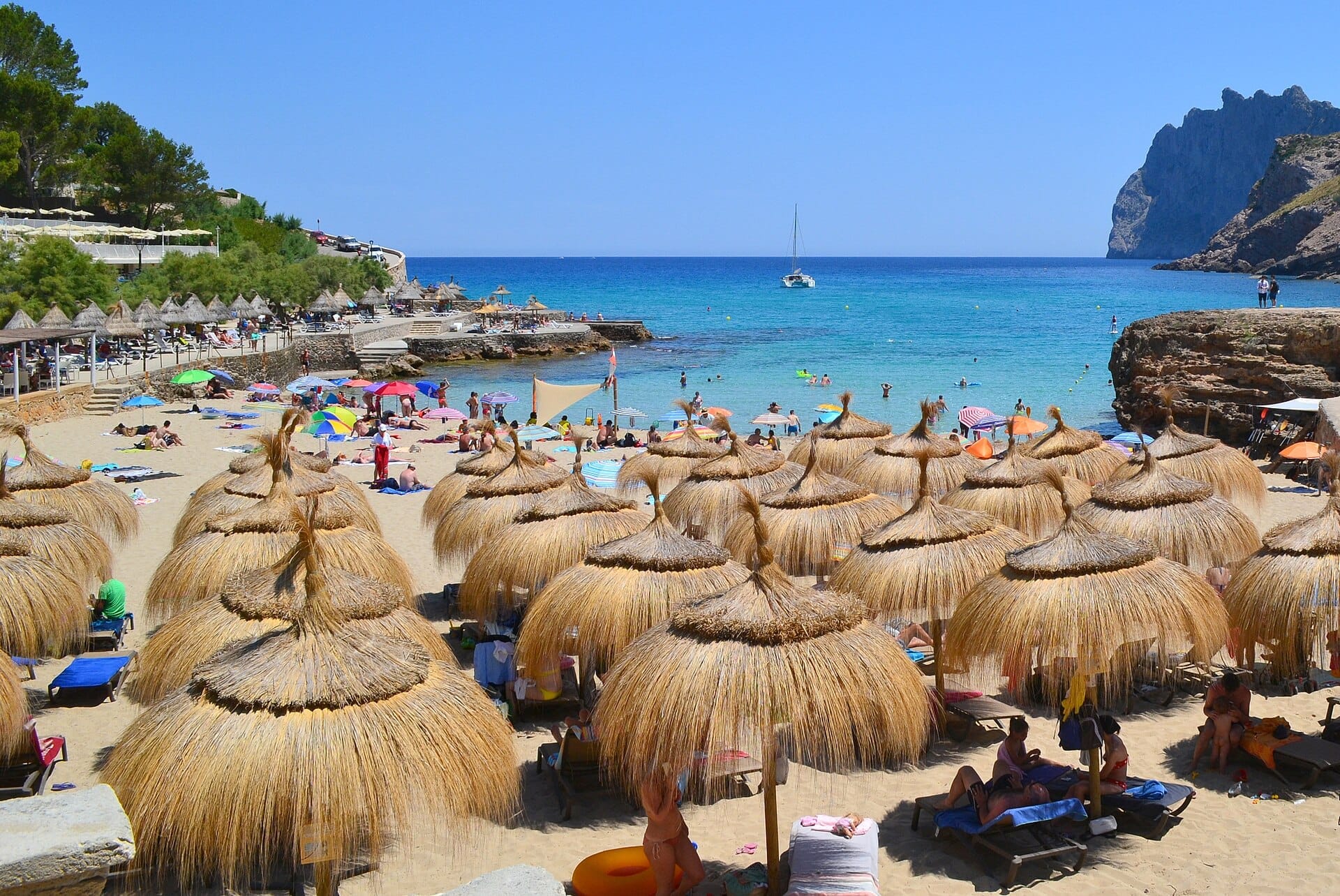 Vista della spiaggia Cala Molins con file di ombrelloni di paglia, bagnanti sulla sabbia dorata, acqua blu cristallina e scogliere ricoperte di pini a Maiorca.