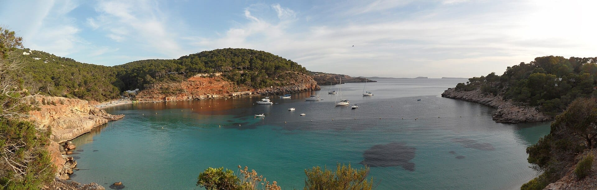 Vista panoramica delle calette Cala Salada e Cala Saladeta a Ibiza, con acque turchesi, barche ancorate, scogliere rocciose e lussureggianti colline verdi.