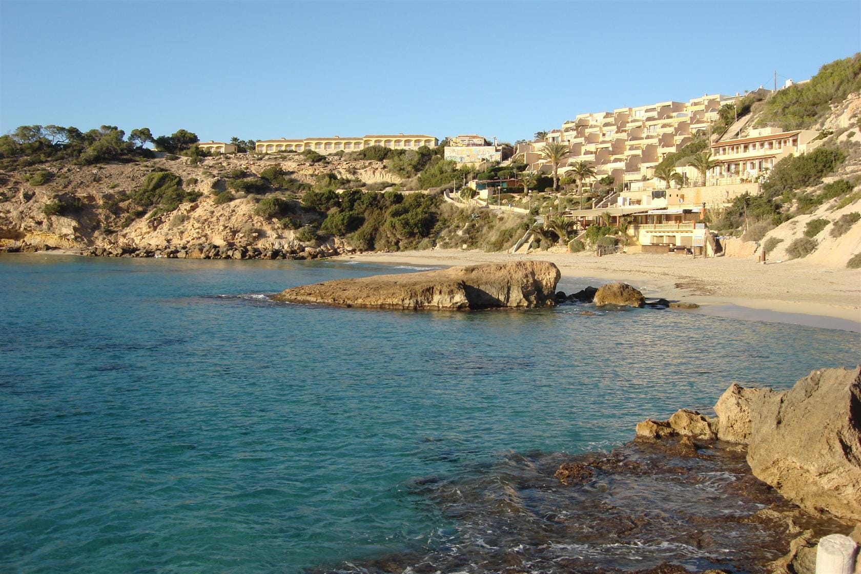 Acque turchesi limpide e sabbia bianca a Cala Tarida, con affioramenti rocciosi e resort sulla pittoresca costa occidentale di Ibiza.