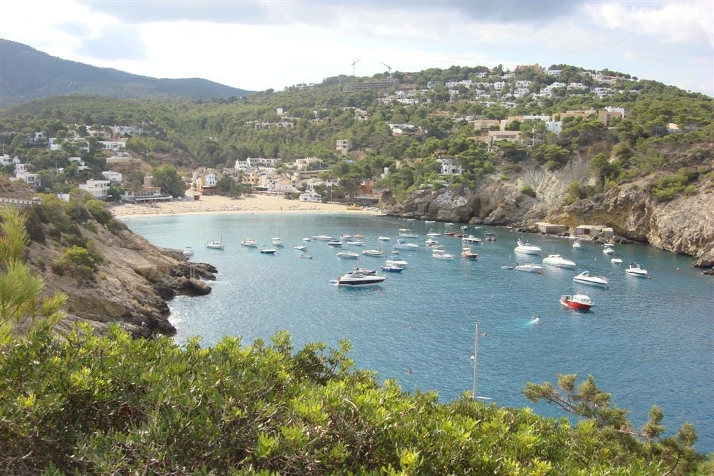 Vista dell'insenatura Cala Vadella a Ibiza con acque turchesi, barche ancorate, spiaggia sabbiosa e ville bianche sul versante collinare circondate da colline verdi.