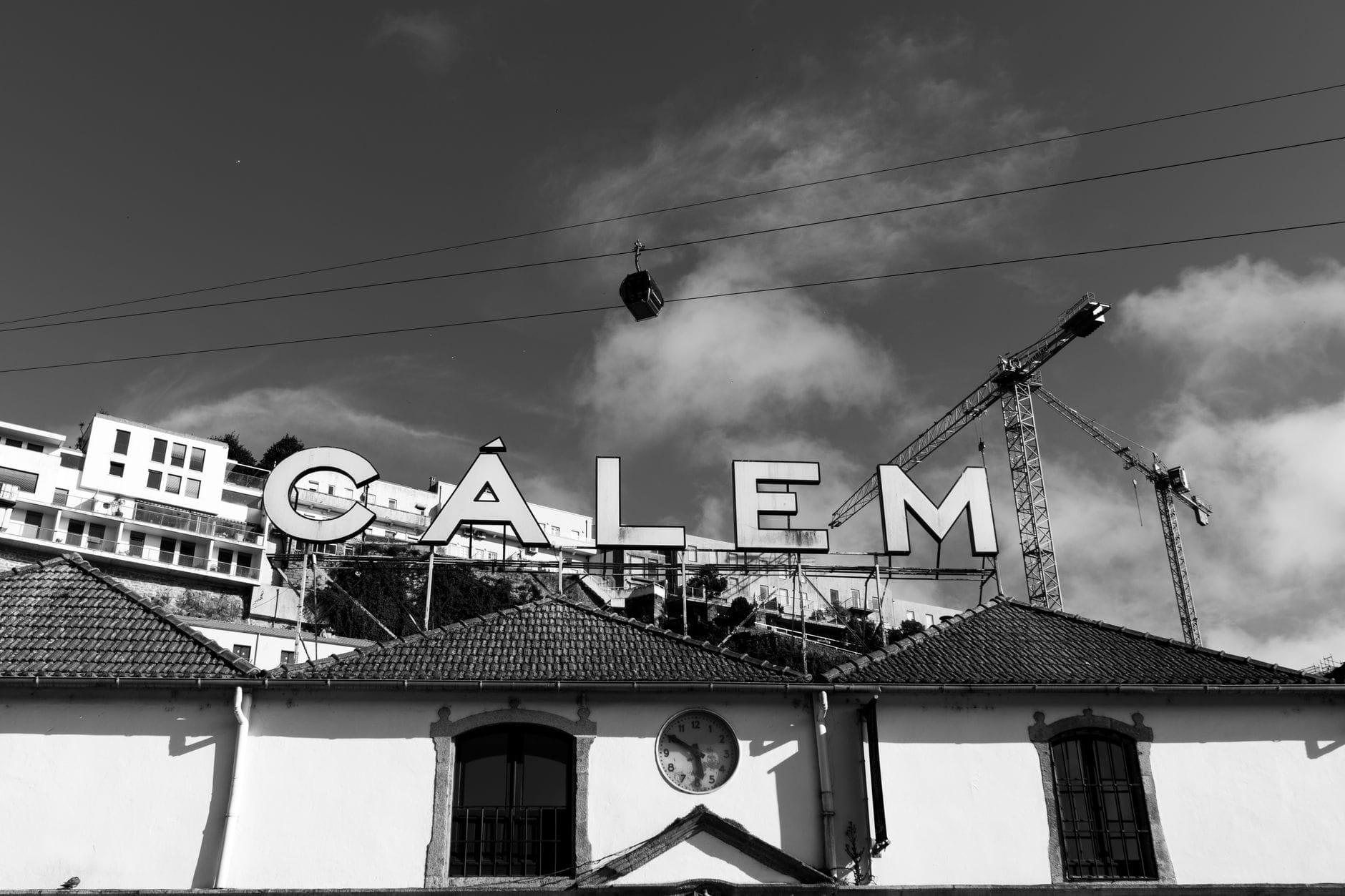 Vista in bianco e nero delle cantine Cálem: l'insegna iconica sul tetto spicca sotto un cielo con funivie e gru a Vila Nova de Gaia.
