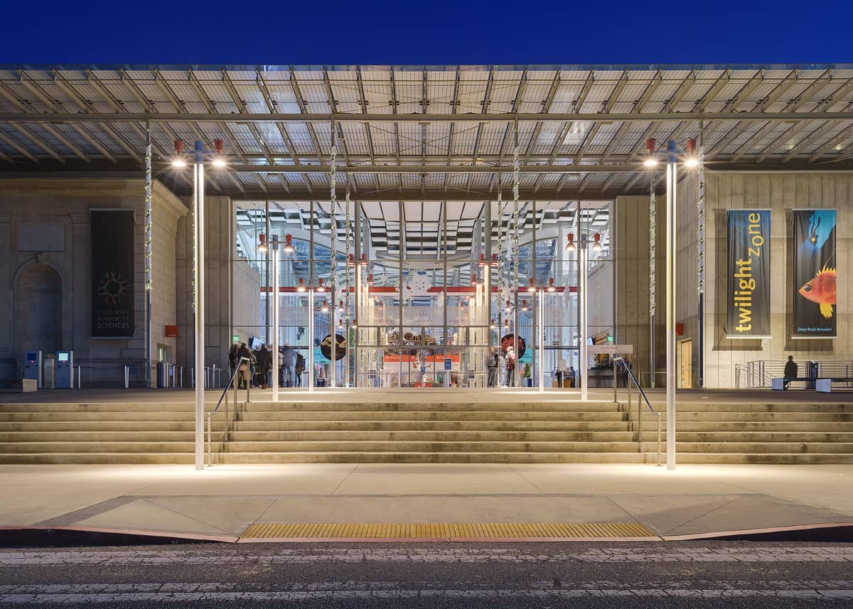 La moderna entrada de cristal y acero de la California Academy of Sciences de noche, con banners y personas visibles en el vestíbulo.